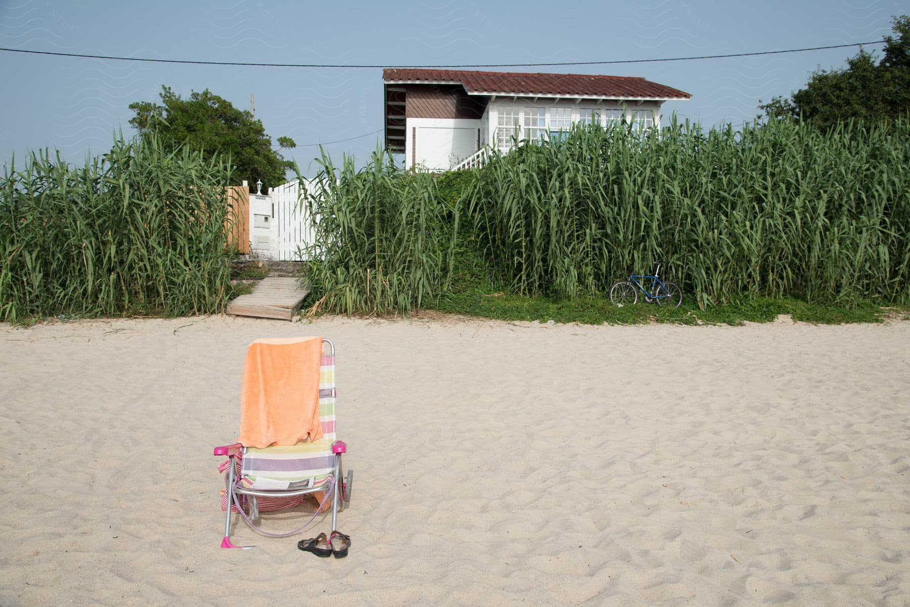 Colorful striped beach chair with an orange towel draped over it on a sandy beach in front of tall grass and a white house.