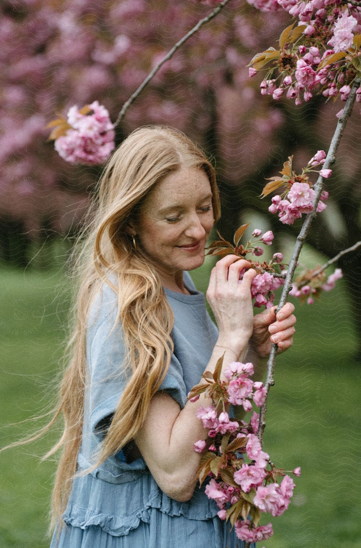 A blonde woman with her eyes closed has a small smile on her face as she hugs a branch full of flowers