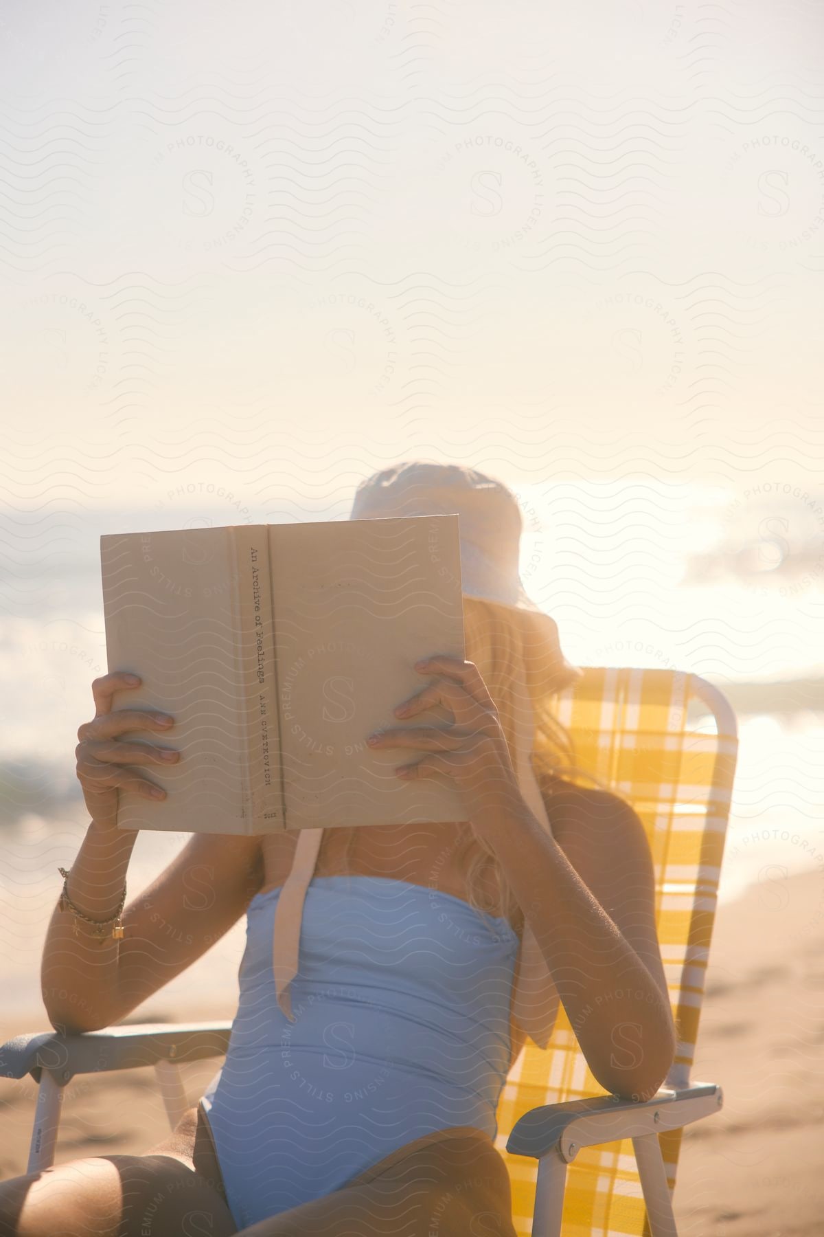 Woman in a blue swimsuit and white hat reading a book while sitting on a yellow-striped beach chair, with the sun shining over the ocean in the background.