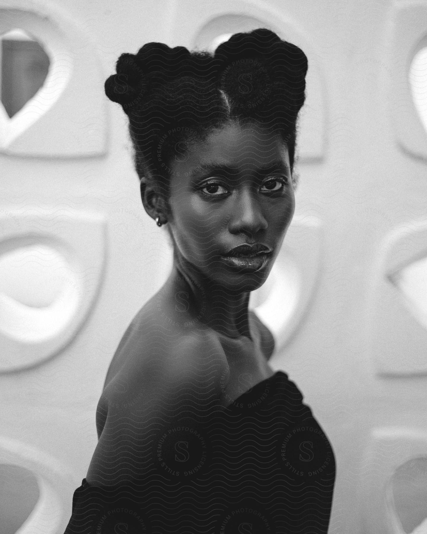 Woman with natural hair styled in two buns, wearing an off-shoulder top, posing against a background with geometric patterns.