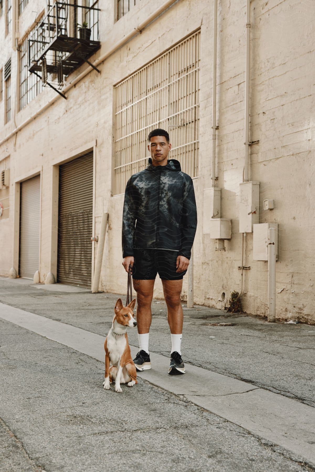 A person in a dark jacket and shorts stands with a dog on a leash in front of a building