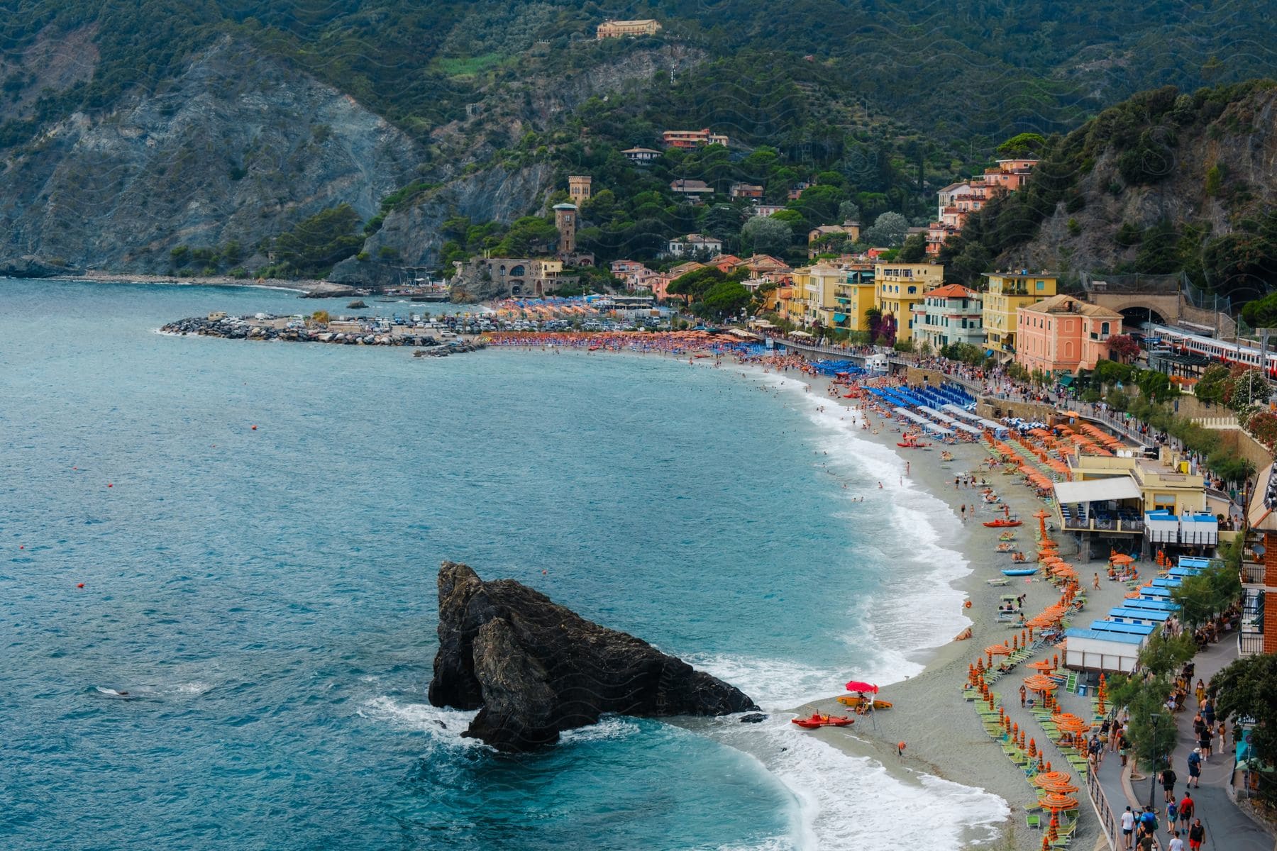 Seaside town with beach umbrellas and sunbathers along a curved shoreline, nestled against a backdrop of green hills and rocky cliffs.