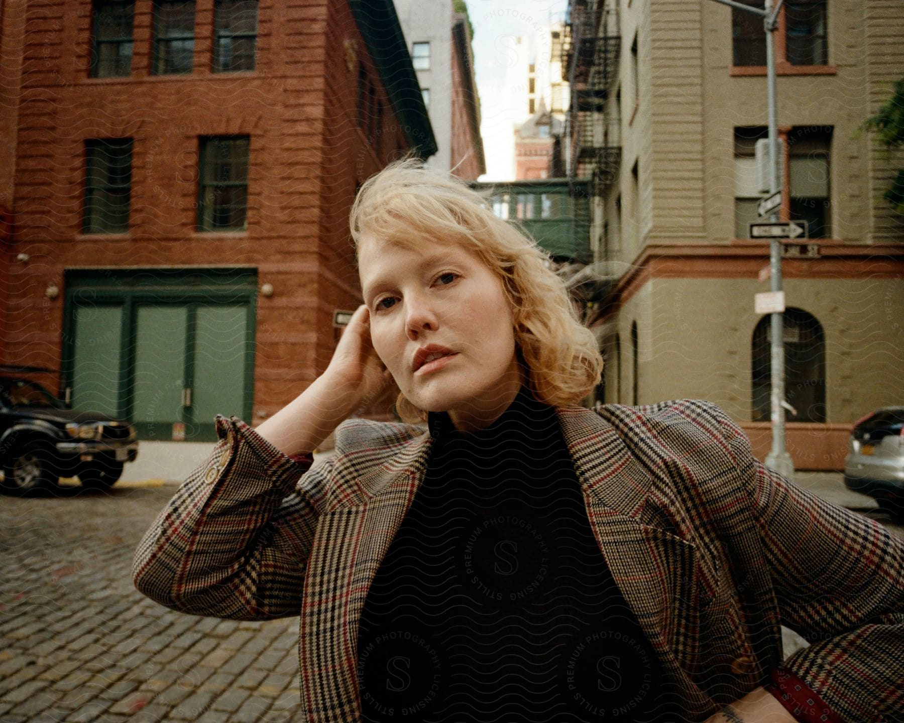 A person with their face in focus on a city street. The person is wearing a checkered jacket and a black blouse, and the scenery includes red brick buildings, a parked car and cobblestone sidewalks.