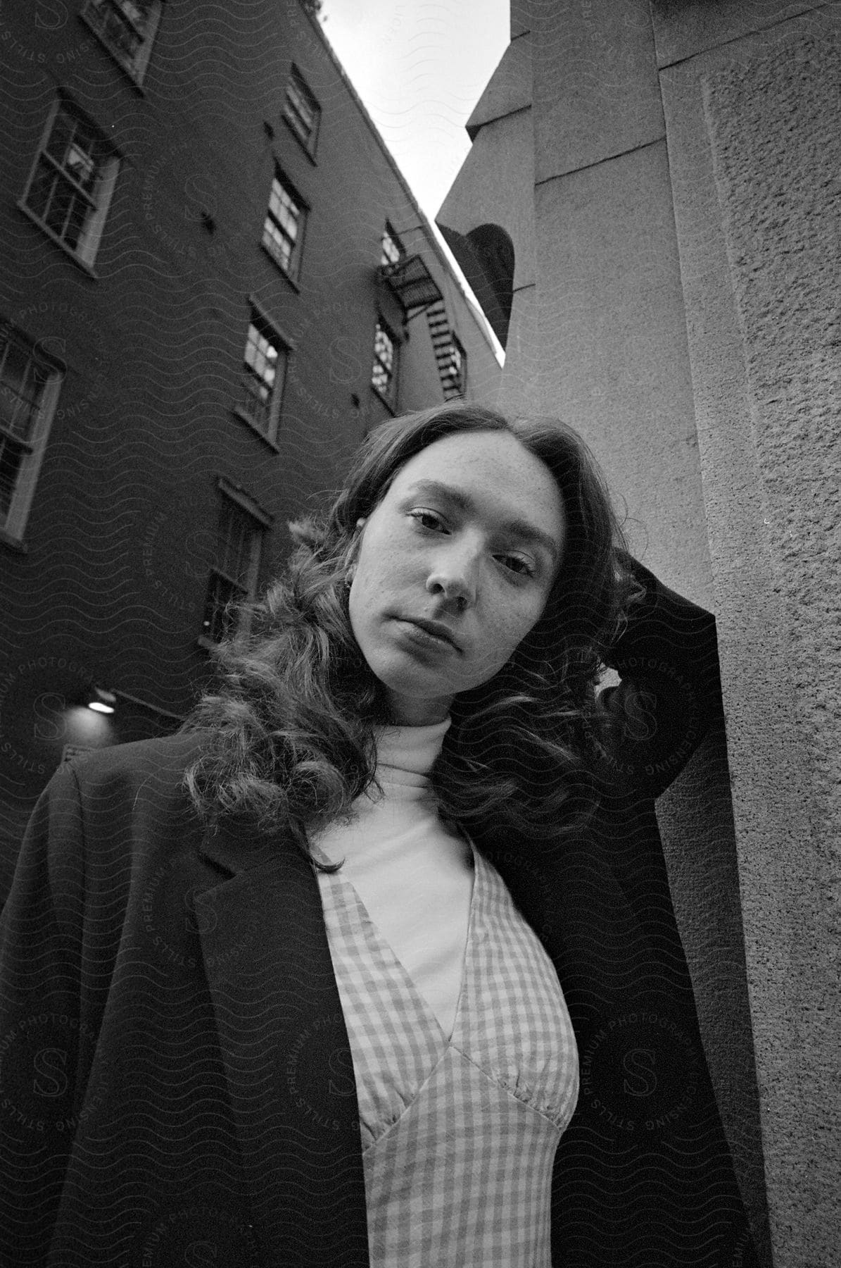 Woman leaning against a building in an urban area with one arm on her head and wearing a black blazer