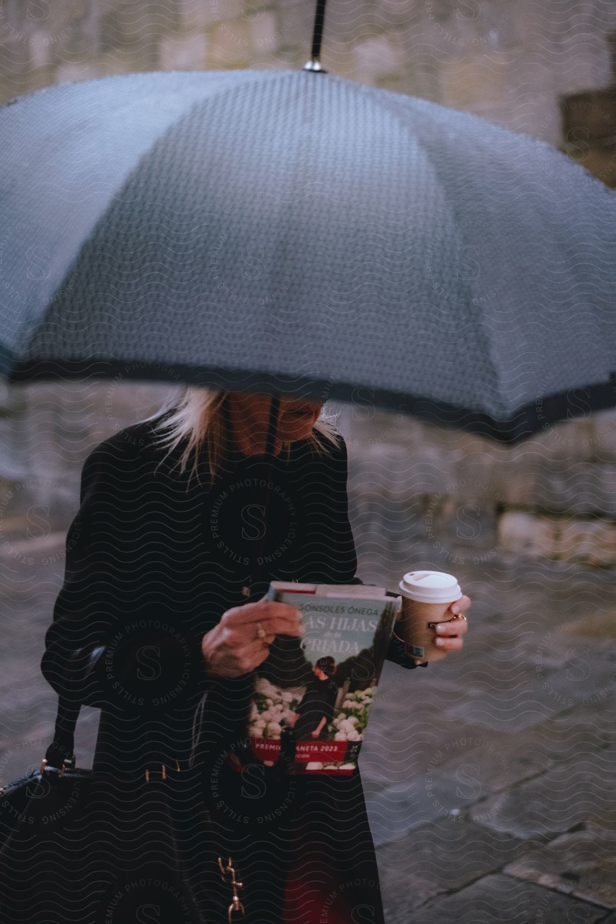 Blonde woman walking on the sidewalk holding an umbrella, book and a cup of coffee