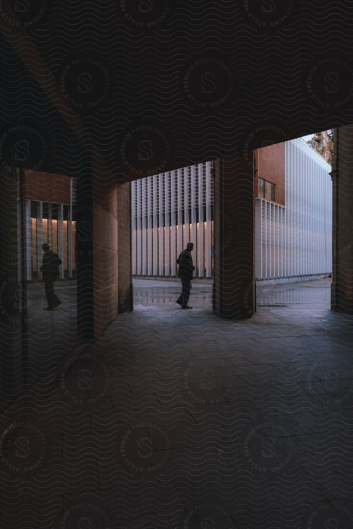 Man walking under the roof of a building as his reflection is seen on the glass door and rain covers the pavement