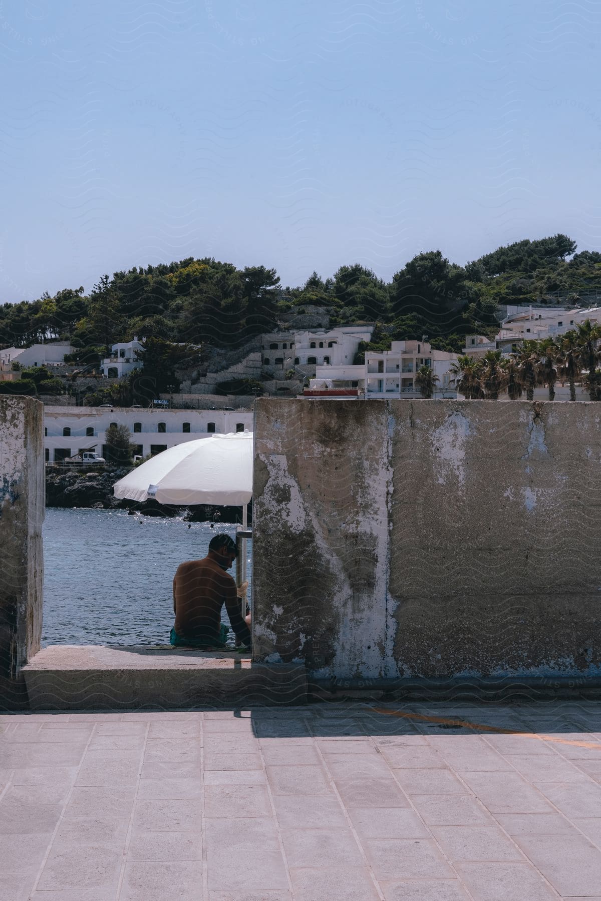 Stock photo of a man sits under a white beach umbrella on a concrete landing on an ocean coast.