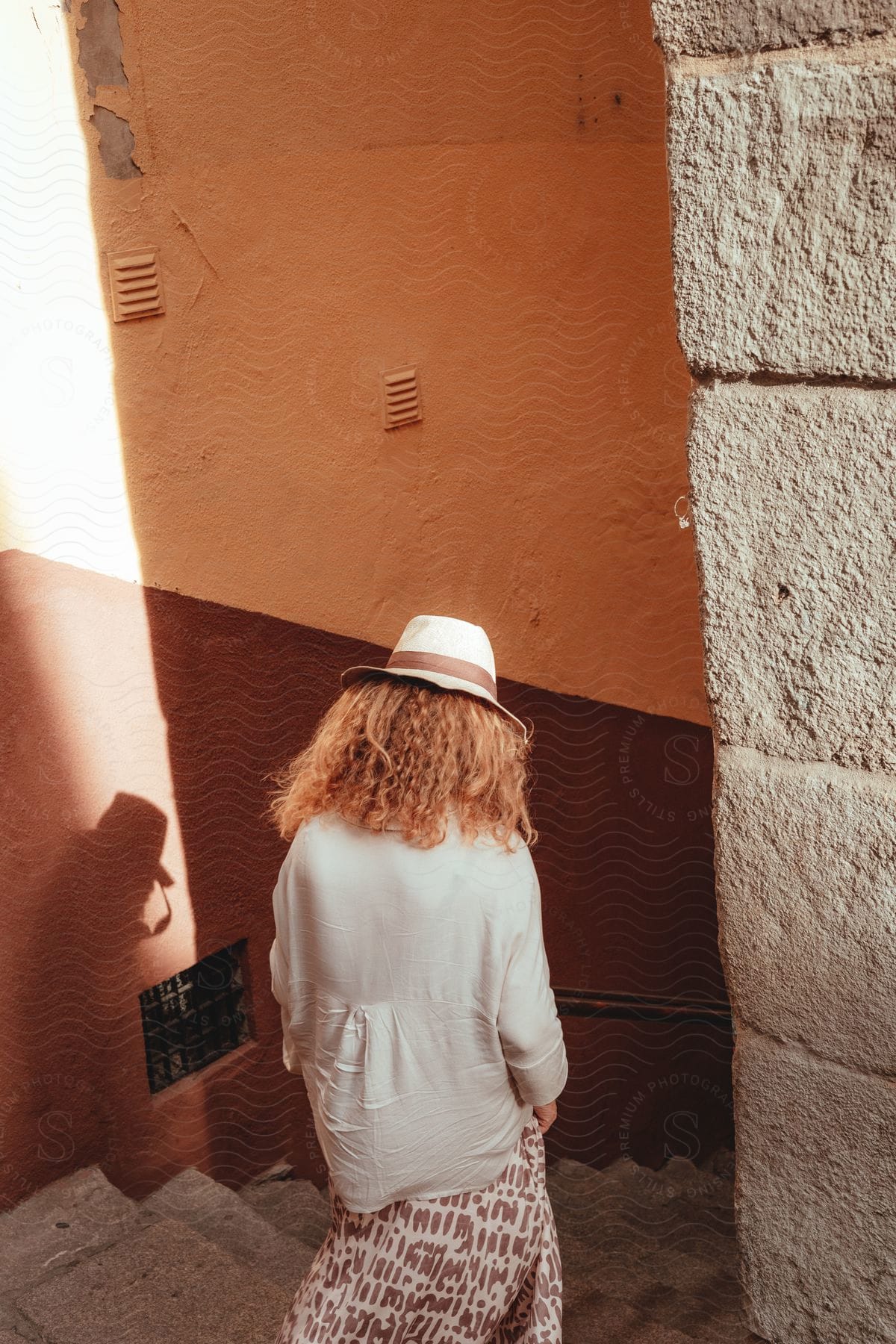 A woman with curly hair wearing a white hat, descends a narrow staircase between orange and brown walls, with sunlight casting her shadow on the wall.