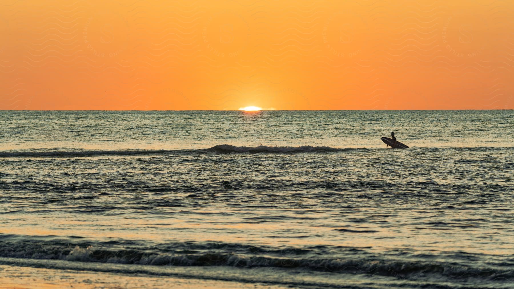 Surfer carrying a surfboard in the ocean at sunset with the sun setting on the horizon and the sky glowing orange.