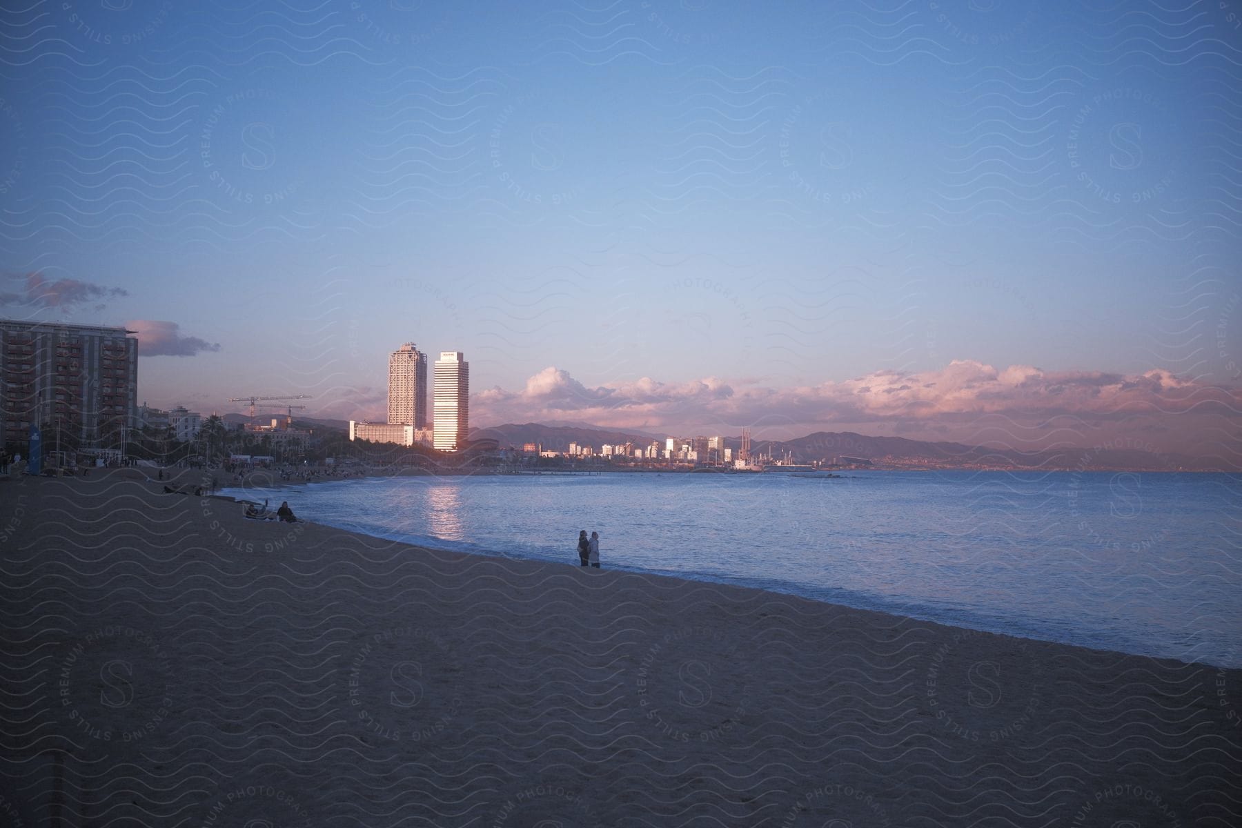 Sunlight reflects off two hotels while people stand together on the beach at sunset.