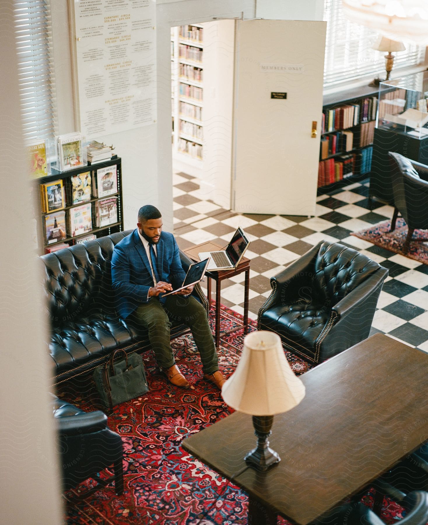 Relaxed on a black sofa in a stylish living room with a black and white checkered floor, a man absorbs himself in the digital world on his tablet.