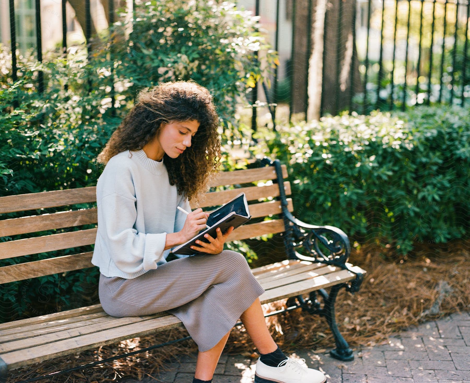 Woman with curly hair sitting on a park bench writing in a notebook with greenery and a fence in the background.