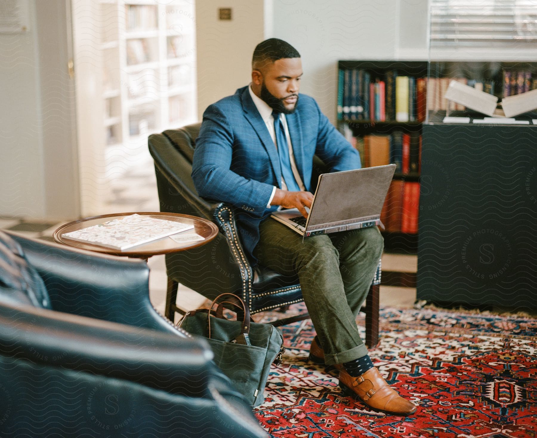 a bearded businessman wearing a blue blazer and tie, green pants, and brown shoes, sitting in a green armchair, and using a laptop in a room with a red carpet.