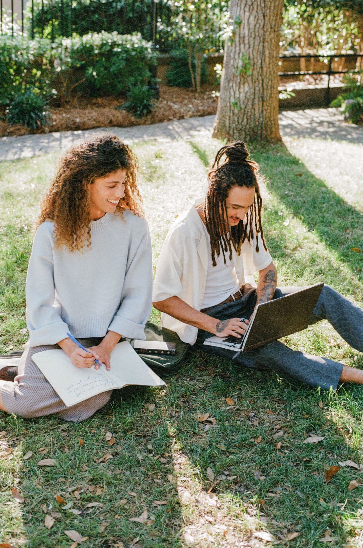 Stock photo of a man with dreadlocks wearing a white top and gray pants, and a woman with curly hair wearing a white shirt and a beige skirt, sitting on a green lawn and using a laptop in a park.