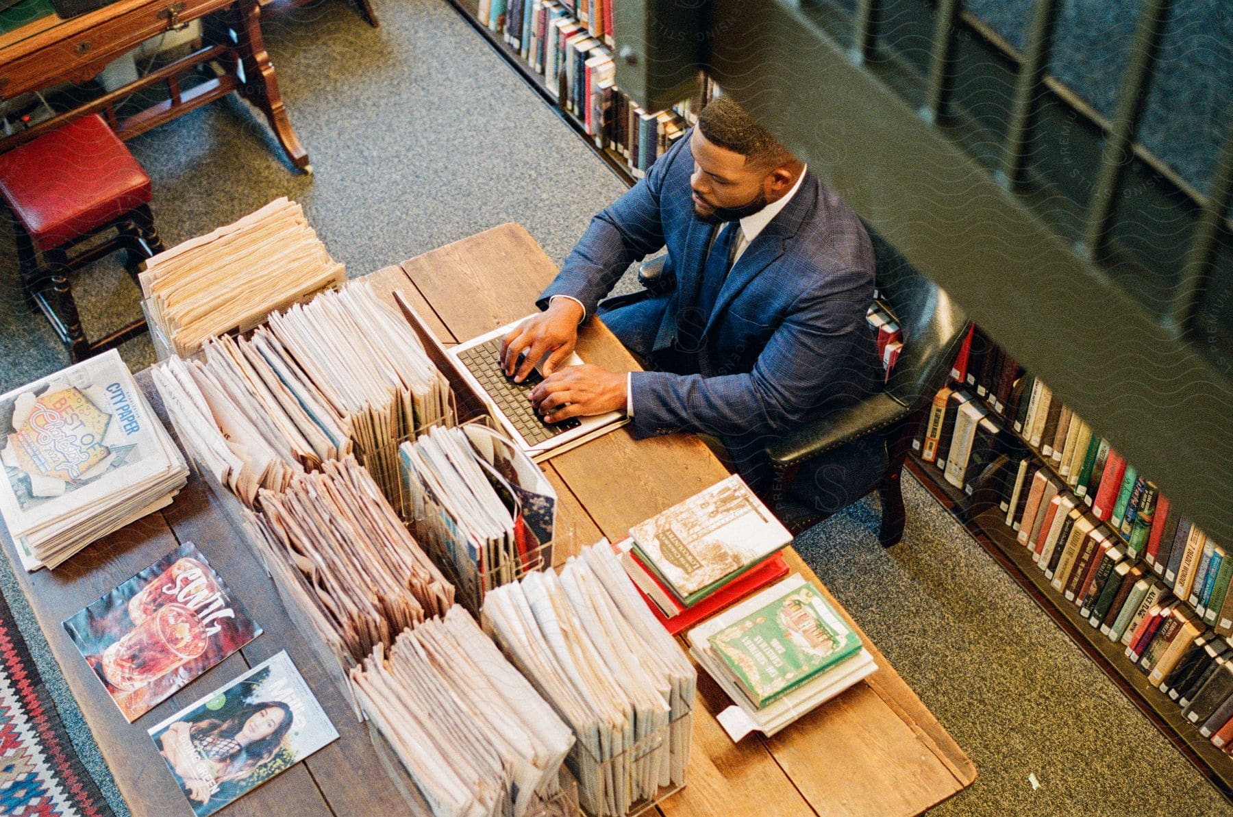Man in a blue suit sitting at a desk typing on his laptop in a library, surrounded by stacks of magazines and books.