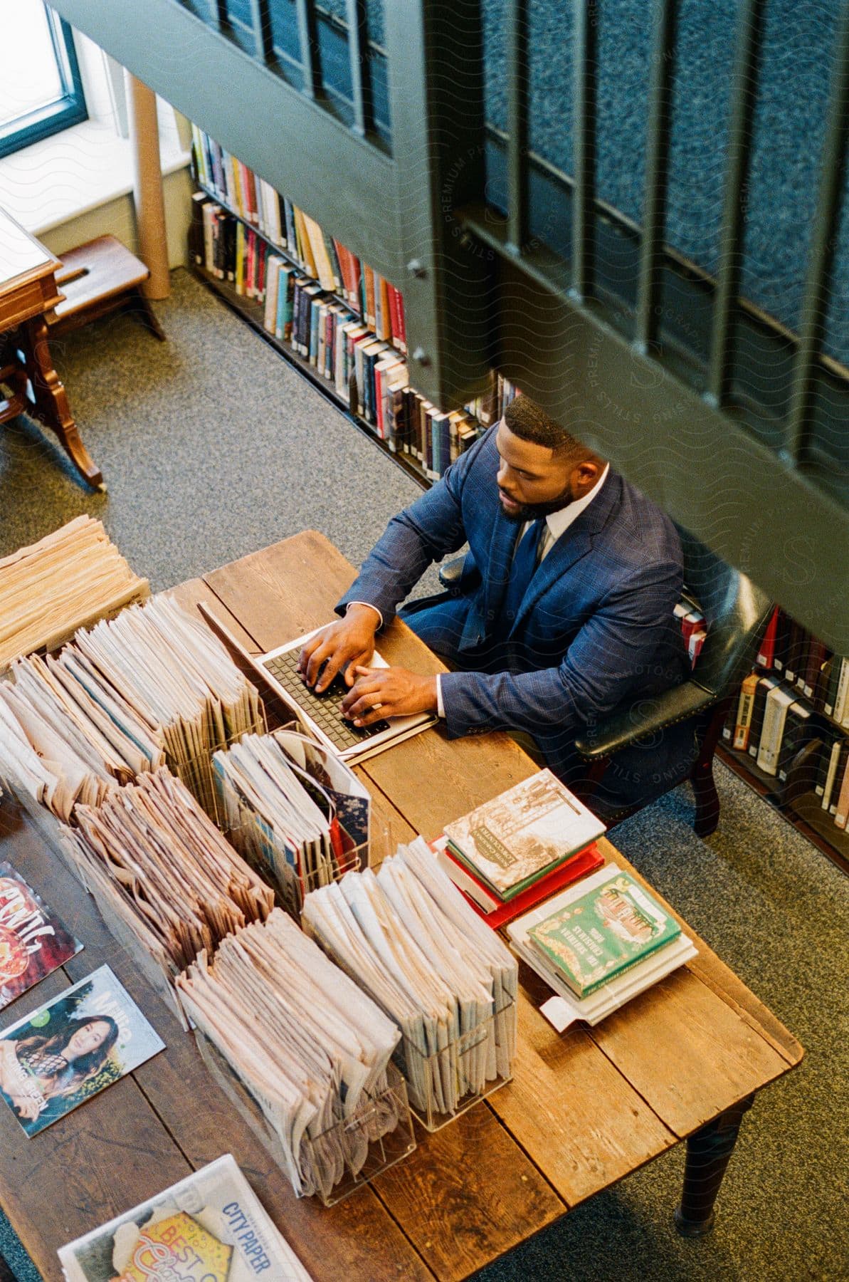 A man in a suit is typing on a laptop that is on a table full of newspapers and books