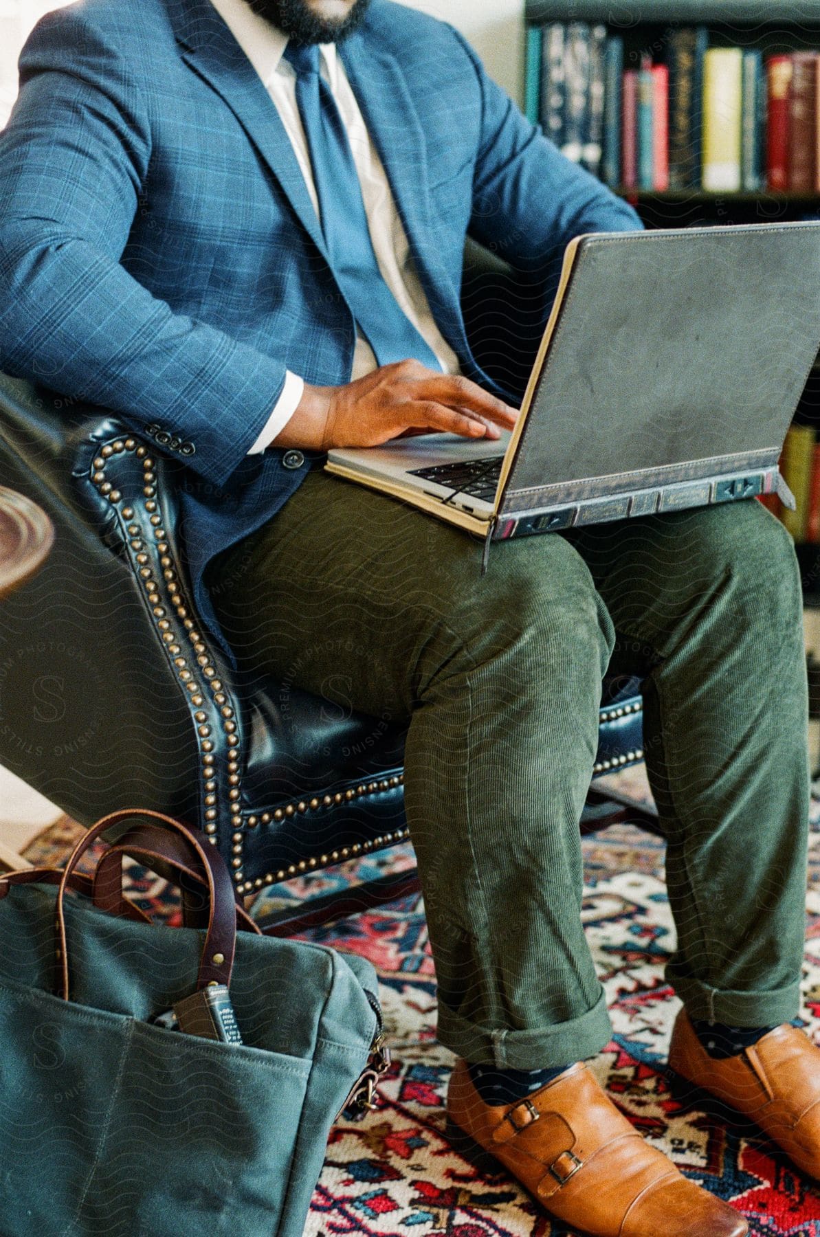 A man wearing a blazer and tie sitting in a leather chair working on his laptop