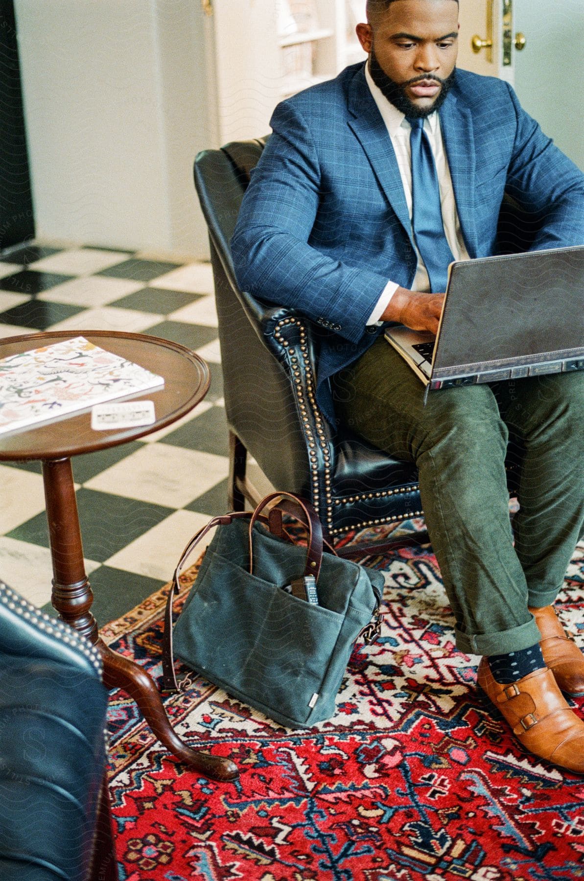 a bearded businessman wearing a blue blazer and tie, green pants, and brown shoes, sitting in a green armchair, and using a laptop in a room with a red carpet.