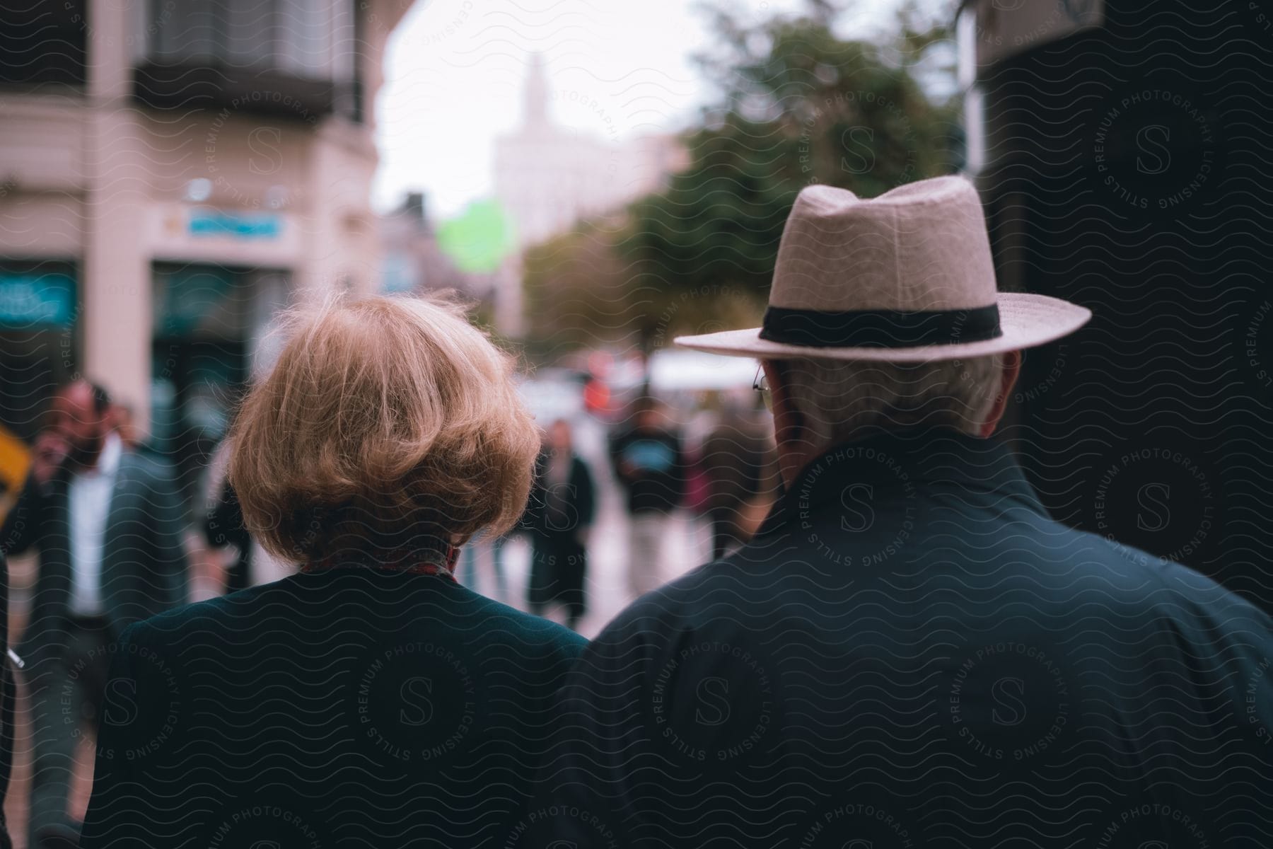 View from behind as a woman and man wearing a hat walk on downtown street among other people