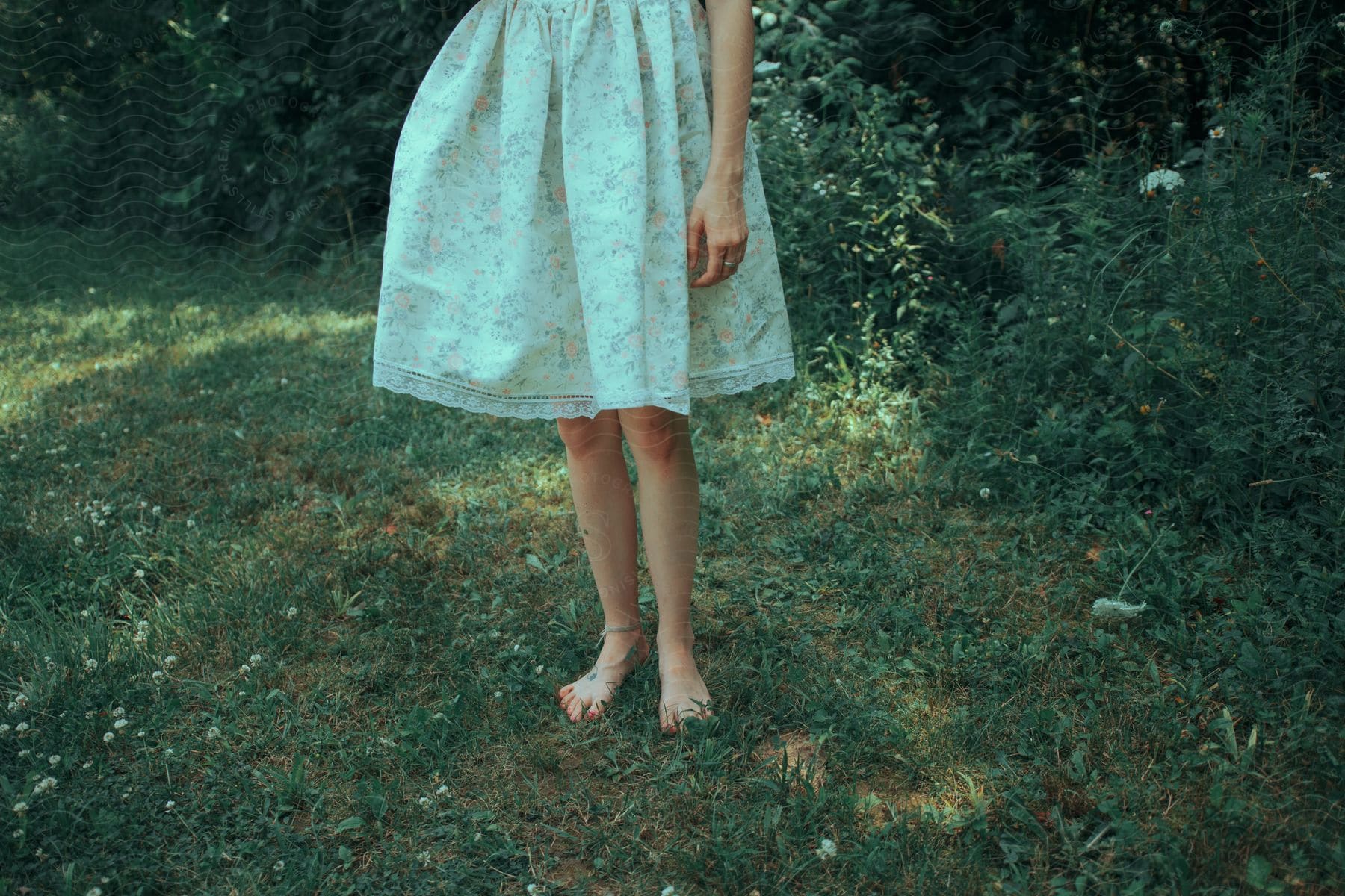 Lower half of a woman wearing a floral dress and an anklet standing barefoot on grass near bushes.