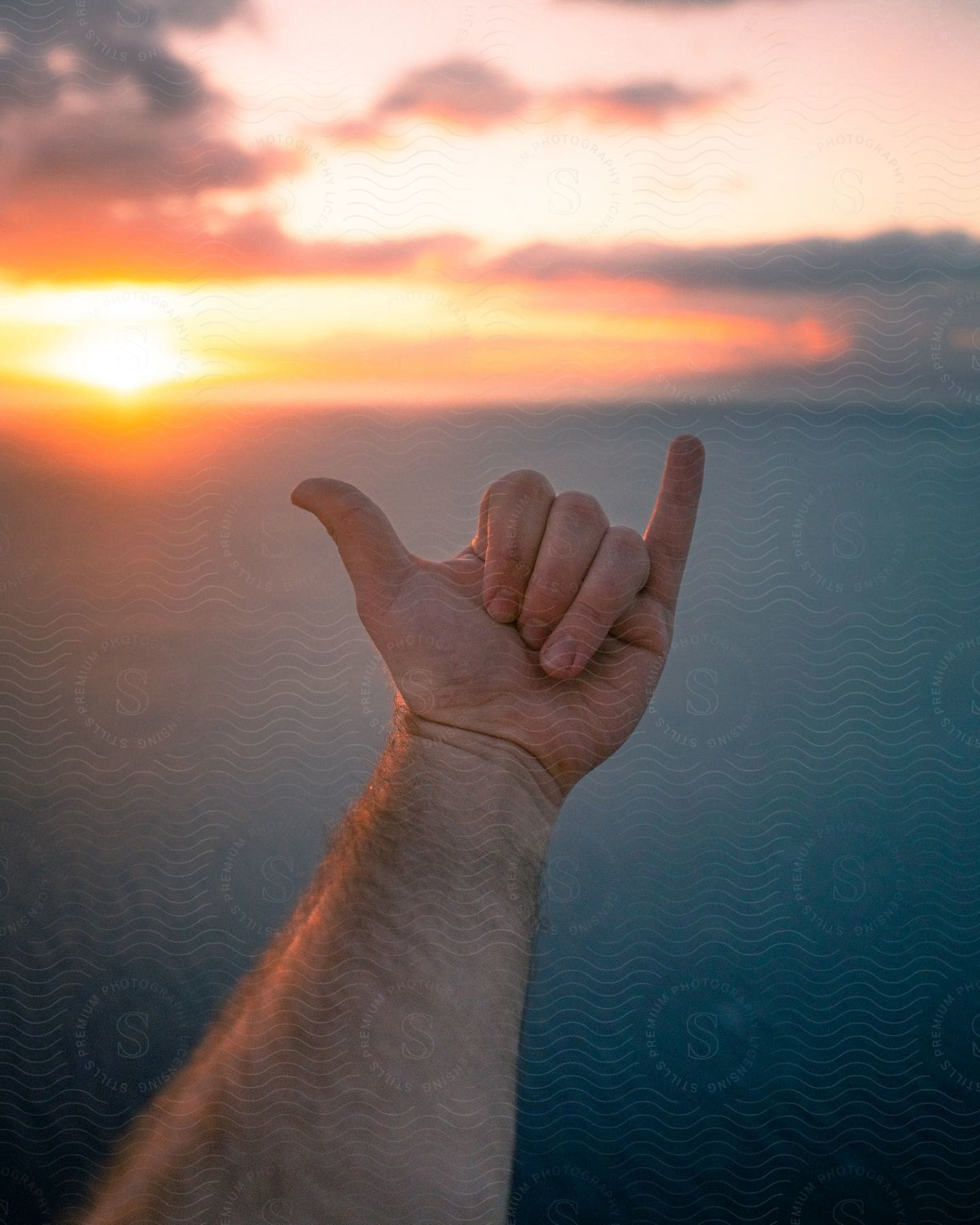 A human hand doing Hang Loose in contrast to a sea and a sky with the sun rising