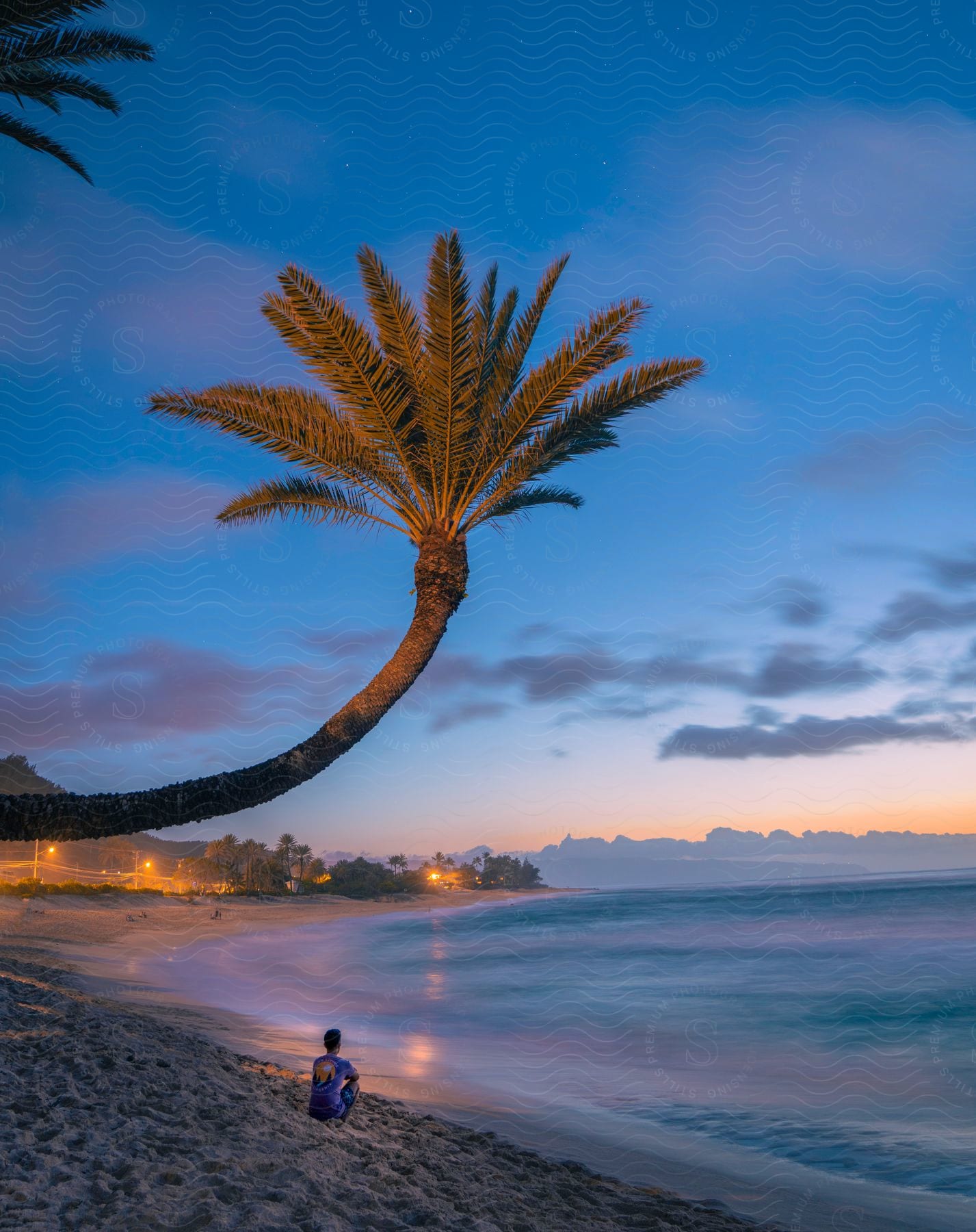 Man sits on the sand on the opposite end of the beach from brightly lit streetlights.