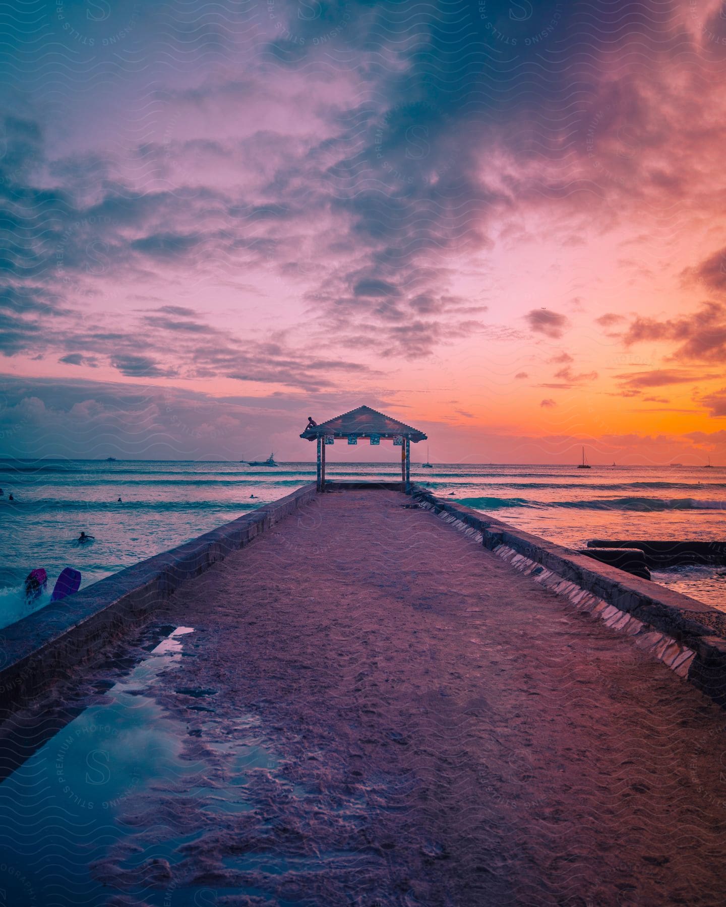 A person sits on the canopy over the pier as people swim in the water and the sun sets on the horizon as boats sail on the water