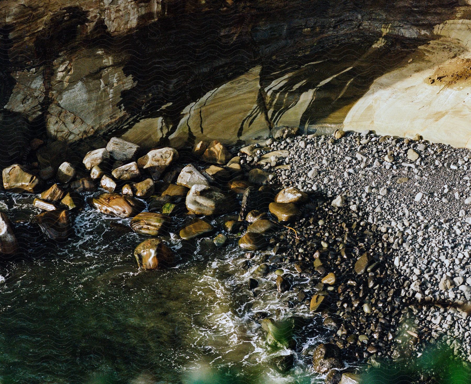 Rocky shoreline of a creek with large boulders and pebbles next to the water, with shadows cast on the rocks and water.