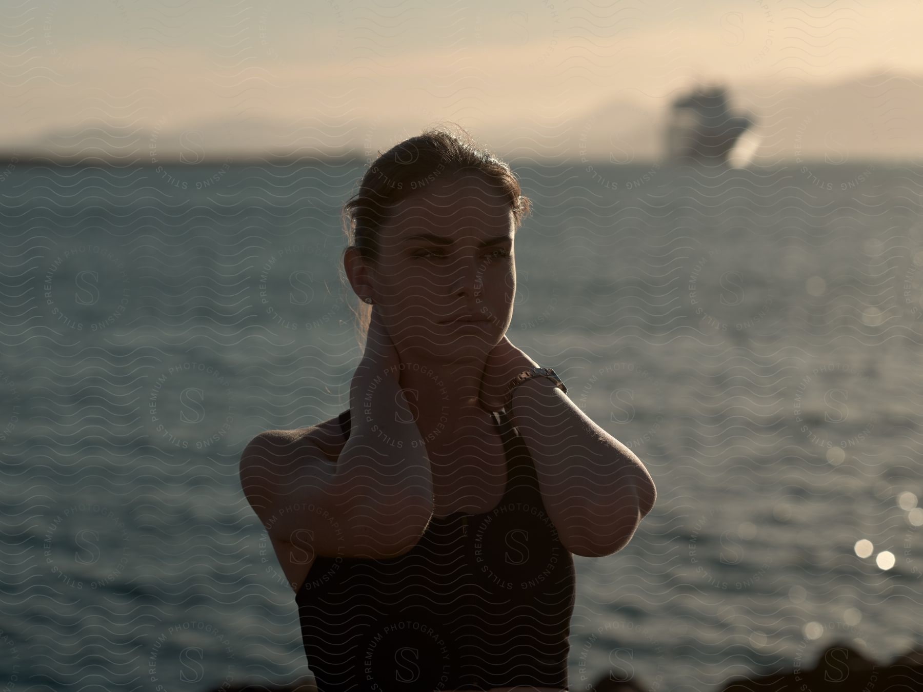 a woman wearing a black top and a wristwatch doing stretches by the sea, with a ship in the background at sunset.