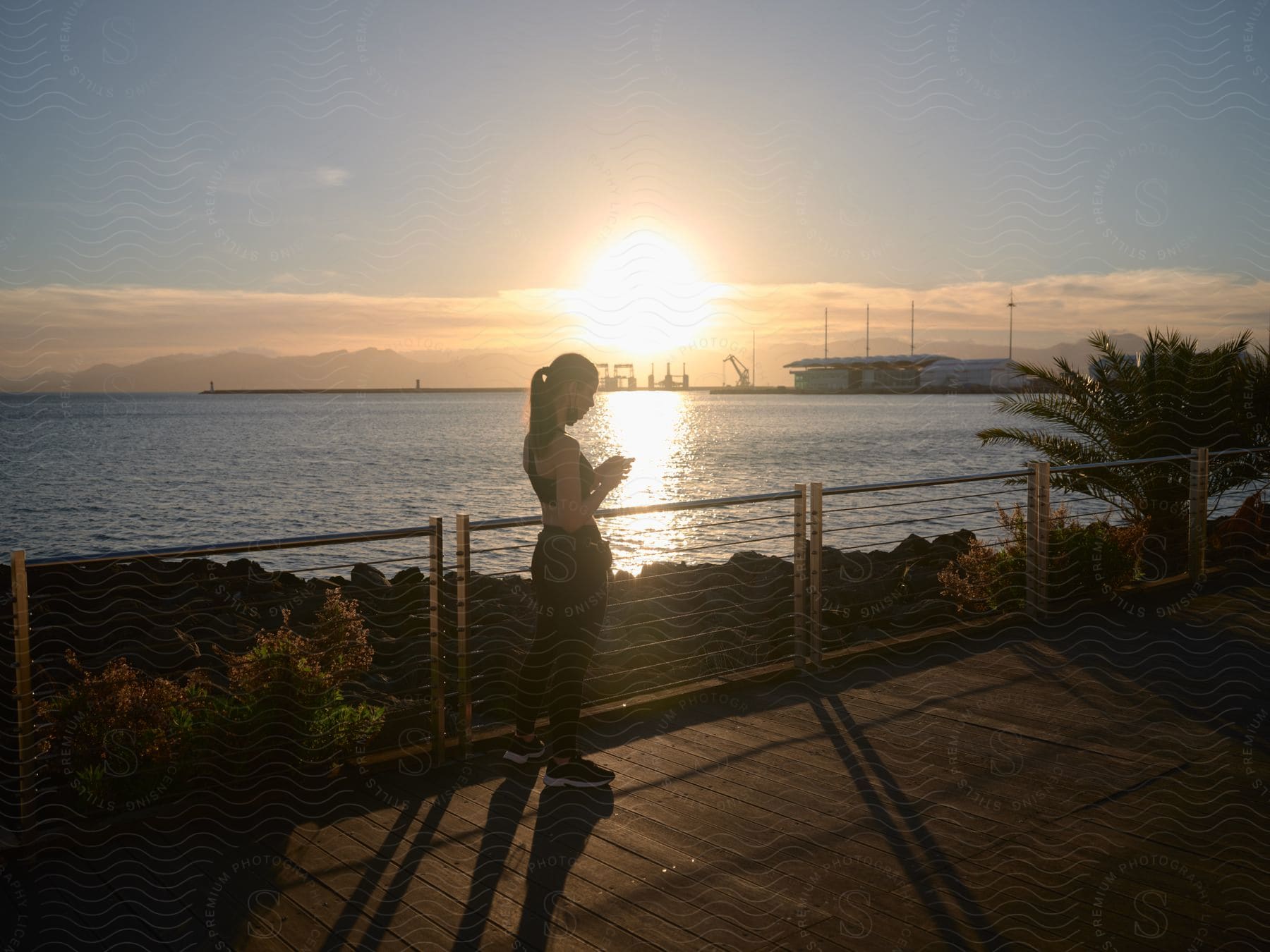 Woman wearing a ponytail and black sports bra stands on a promenade near the sea at sunset