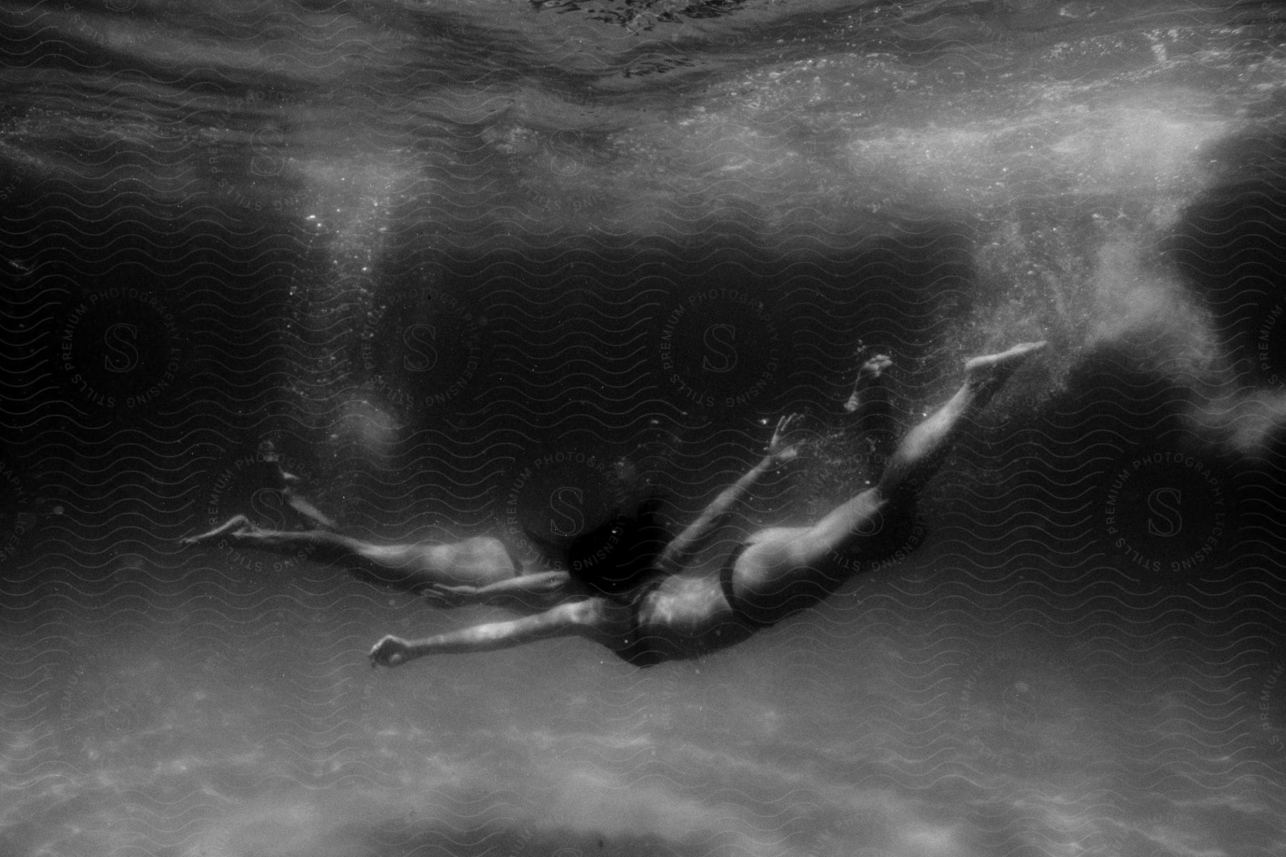 Submerged in the crystal-clear water of a swimming pool, two women clad in black bikinis gracefully plunge into its depths.