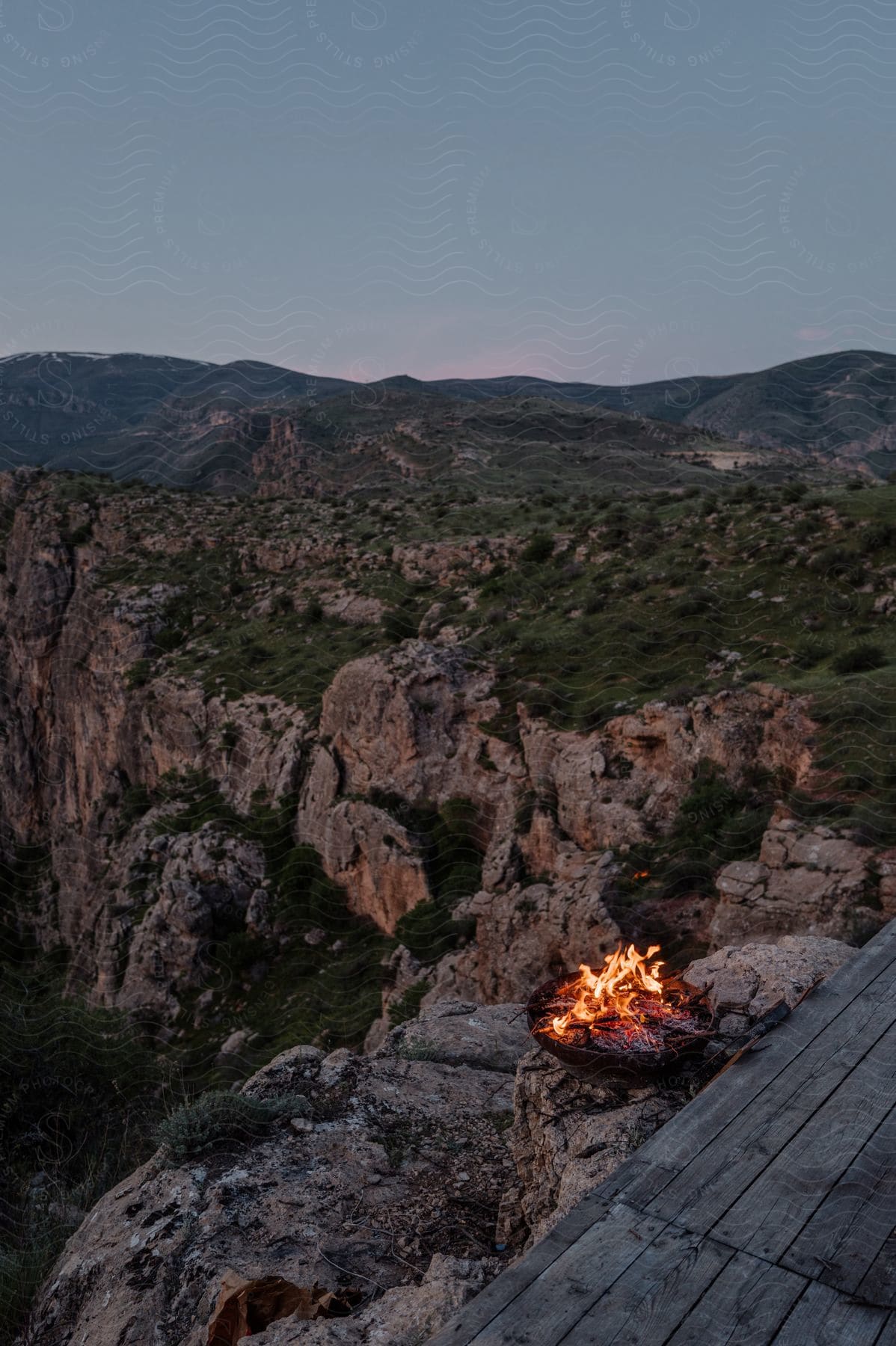 A small campfire set on a wooden platform, overlooking a scenic, rugged mountain landscape during twilight.