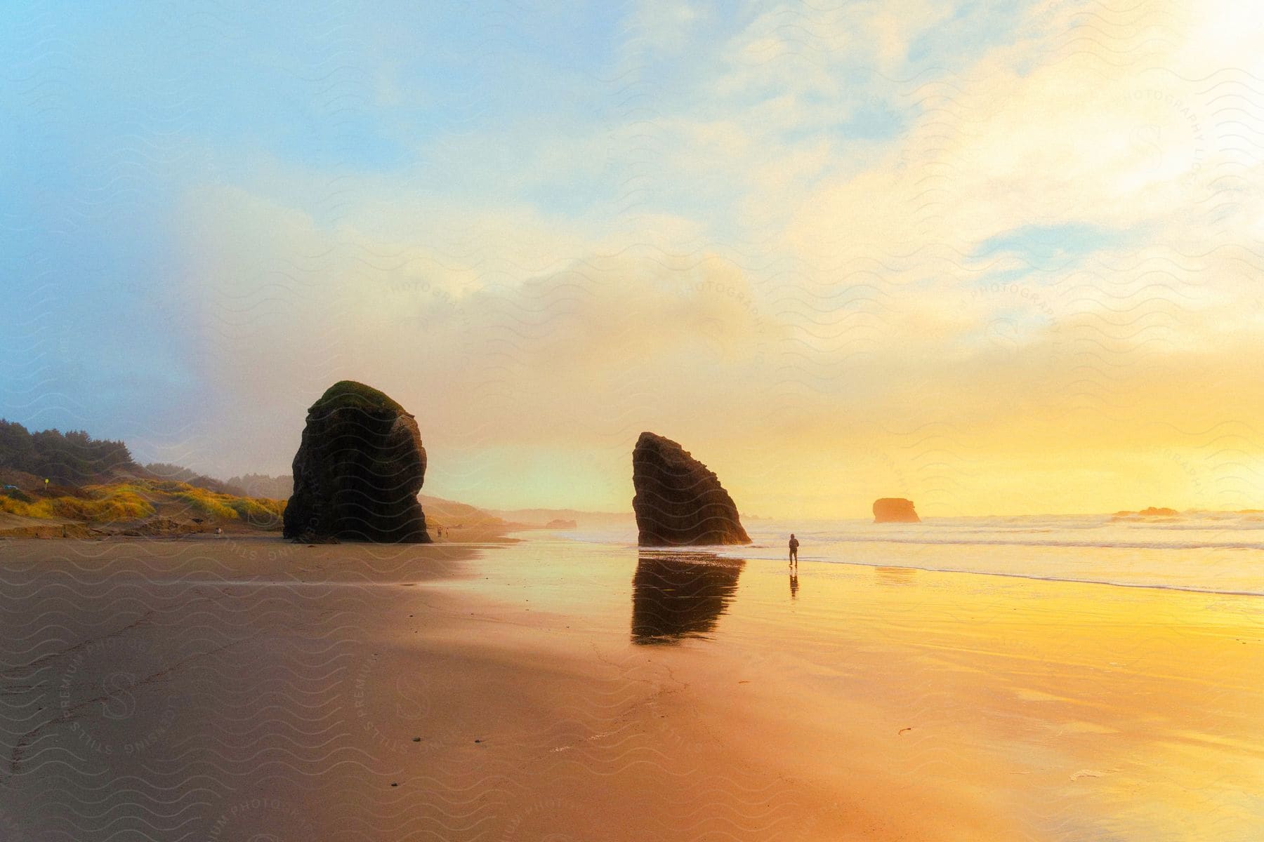 Silhouette of a person walking on the beach among tall rock formations on the coast as sunlight reflects on the clouds and the water