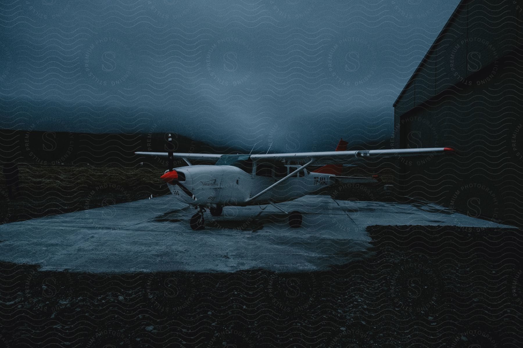 A solitary sensa plane rests under a cloudy night sky on a mountain airport's tarmac, bathed in the soft glow of nearby hangar lights.