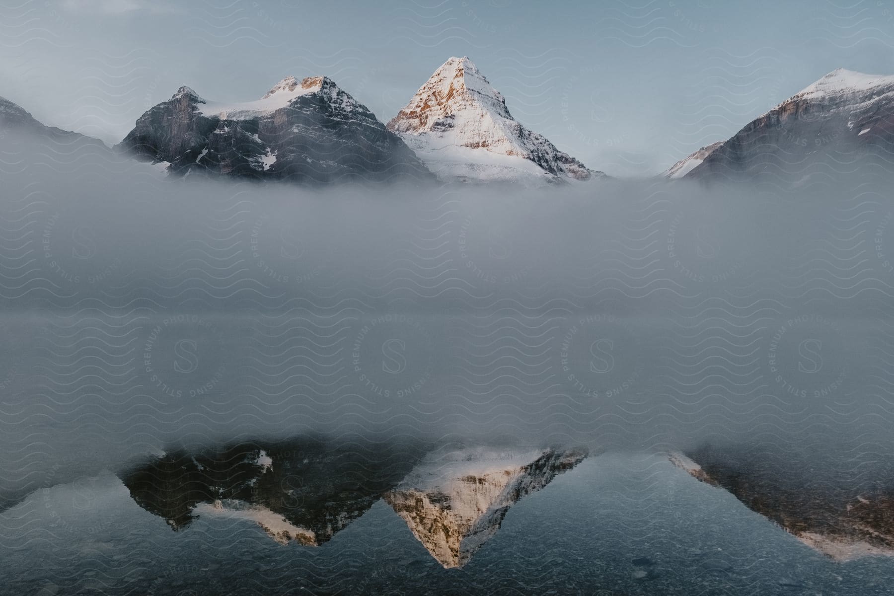 Snow-capped mountain peaks emerging from a dense layer of mist, perfectly reflected in the still waters of a serene lake.
