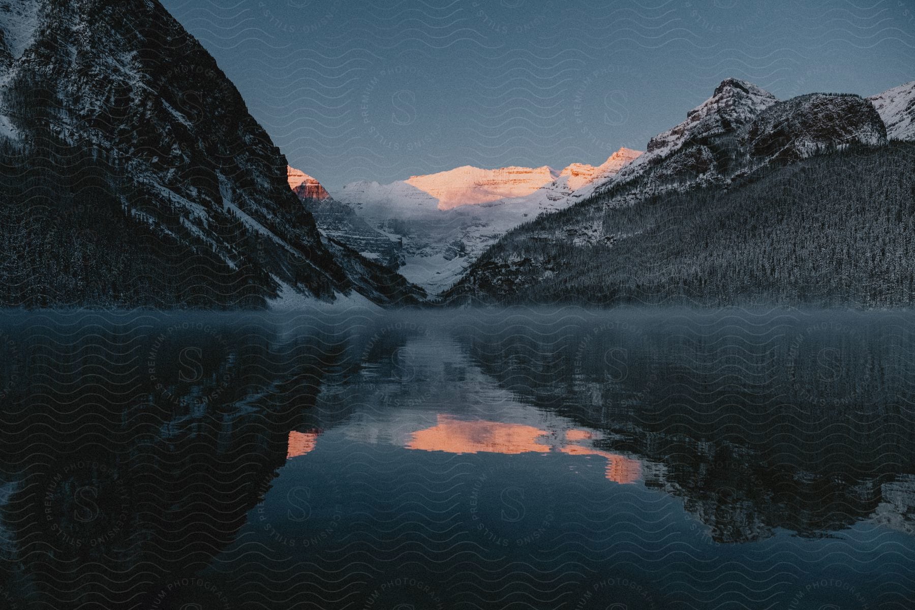 Natural landscape of a calm lake reflecting mountains with snow around in a dark early morning sky