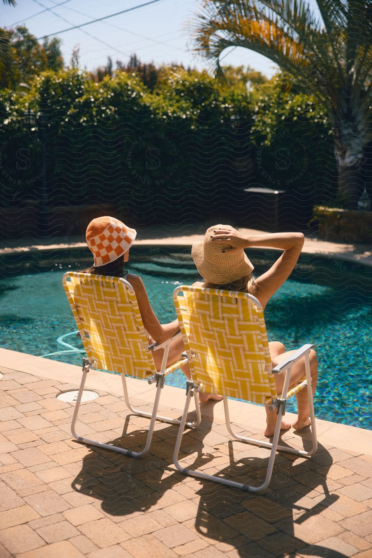 Two women are sitting on beach chairs in front of the pool and both are wearing sun hats.