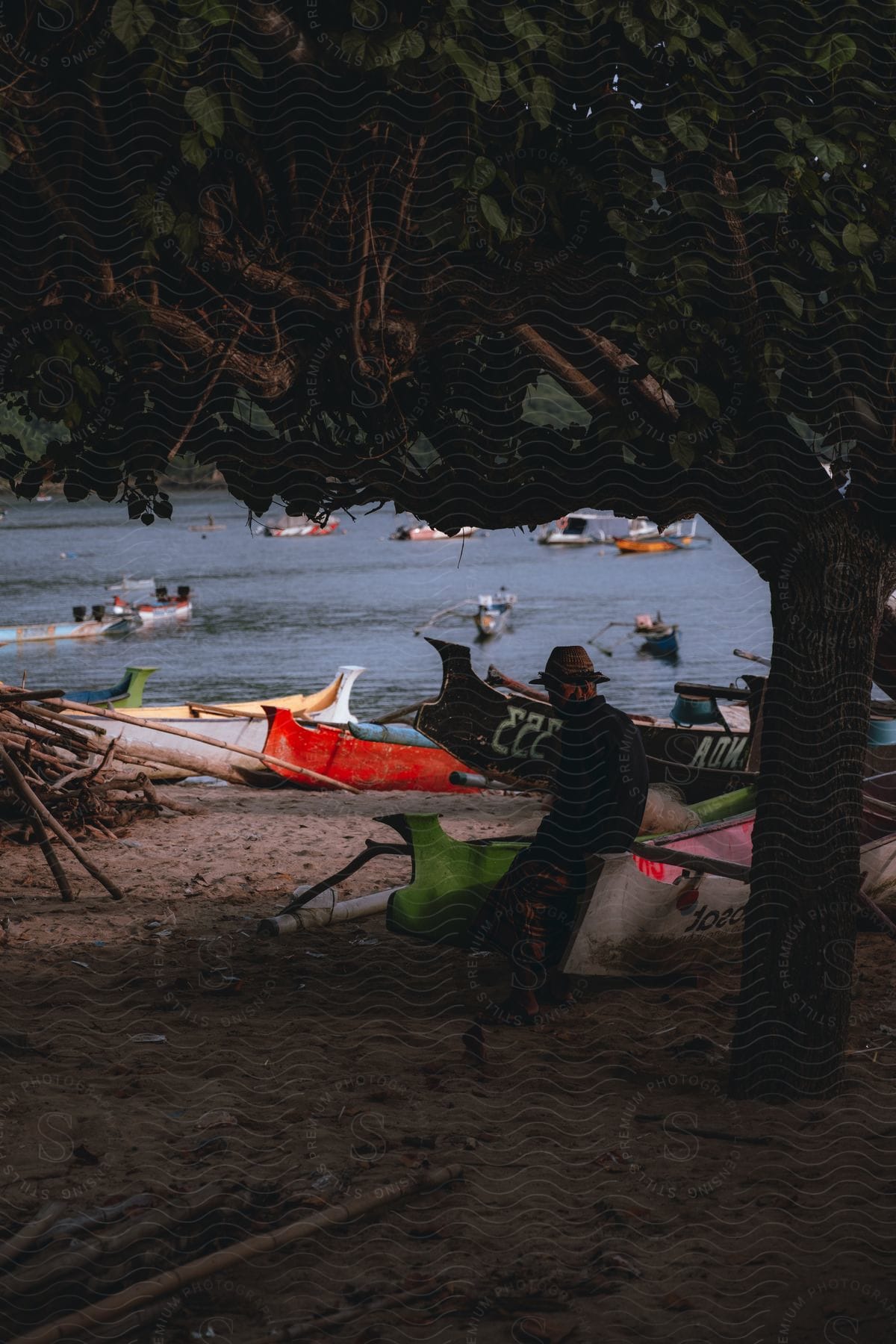 A person sitting next to colorful boats on the sand, overlooking a body of water where other boats float in the distance.