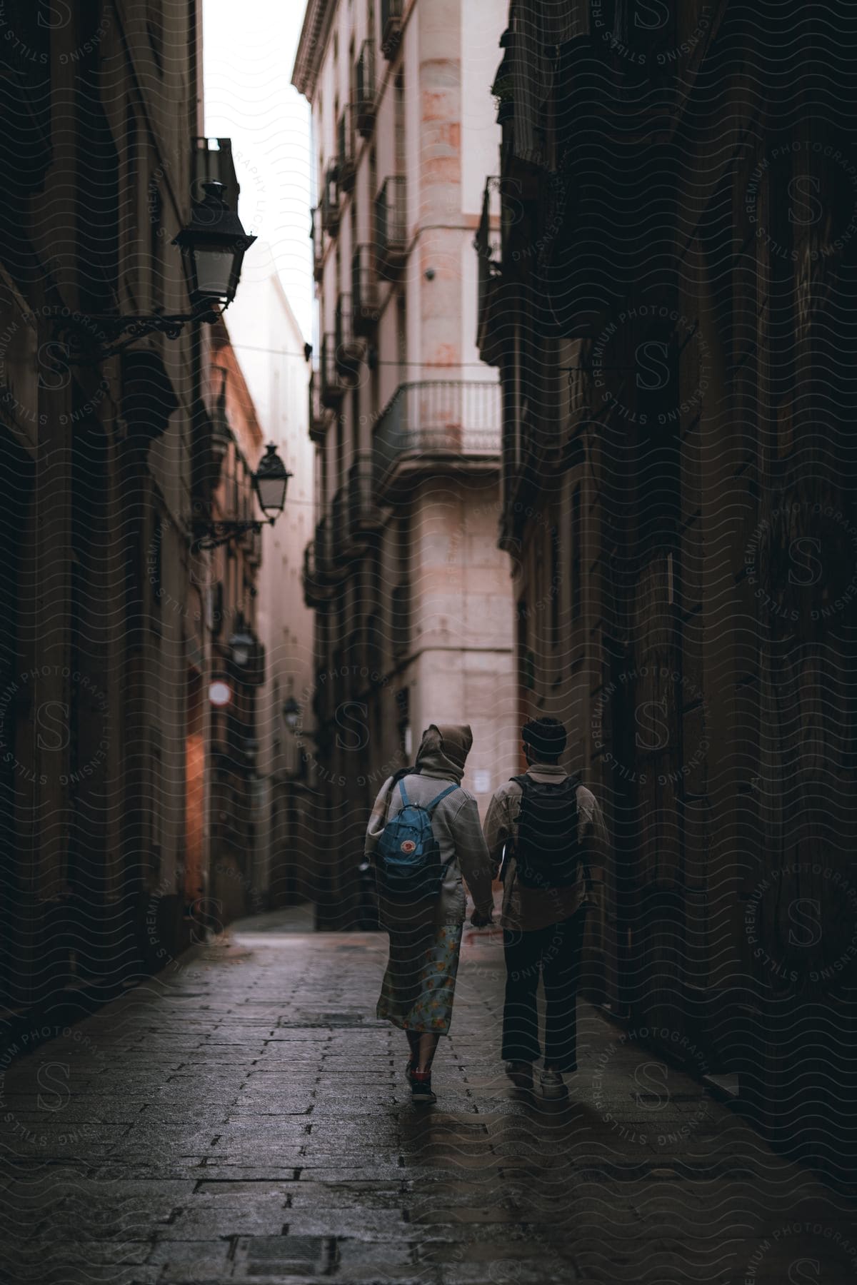 Two people holding hands walking along a narrow paved street between old buildings.