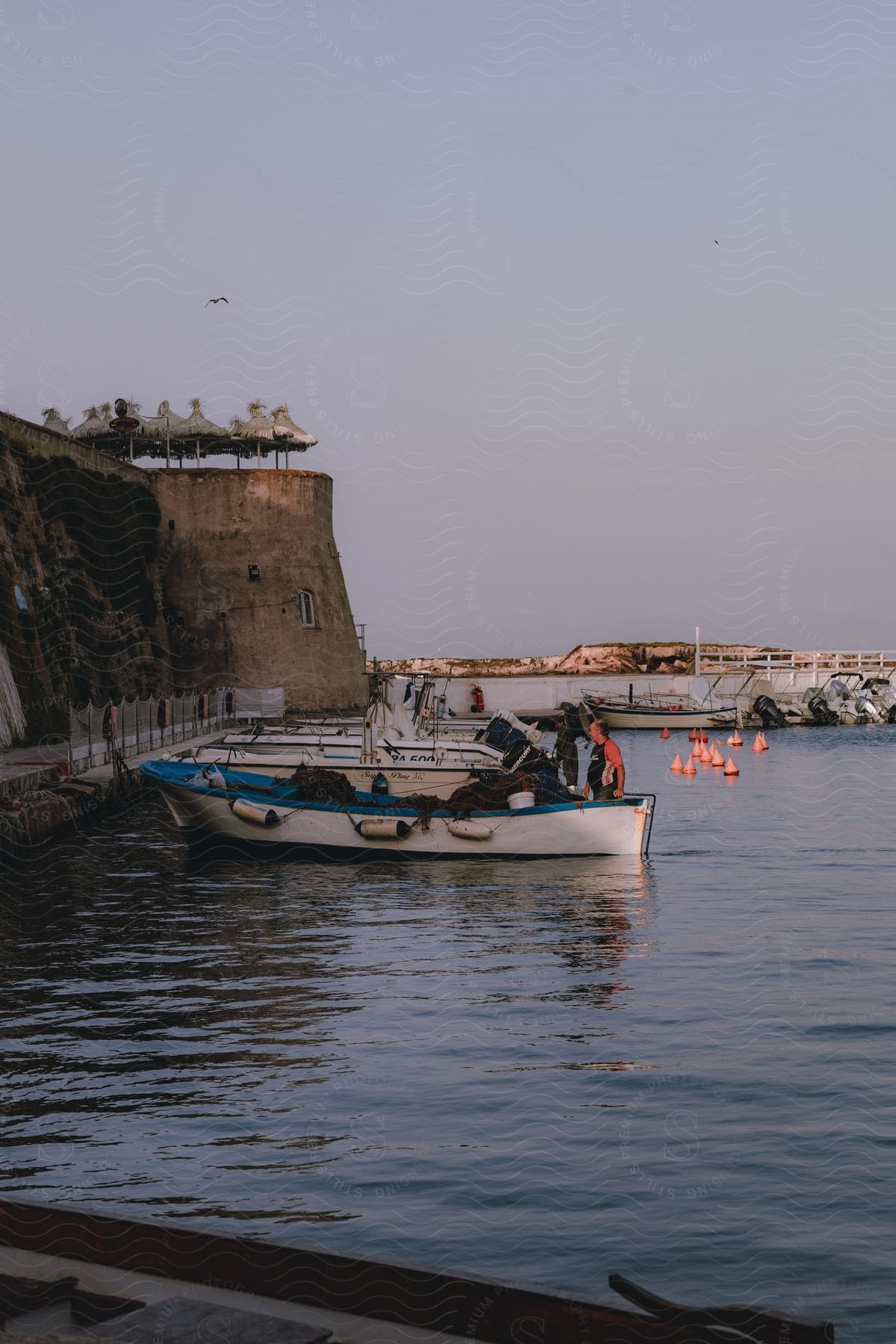 A fisherman working on a small boat moored at a harbor with a large cliff and a structure on top, under a pink sky.
