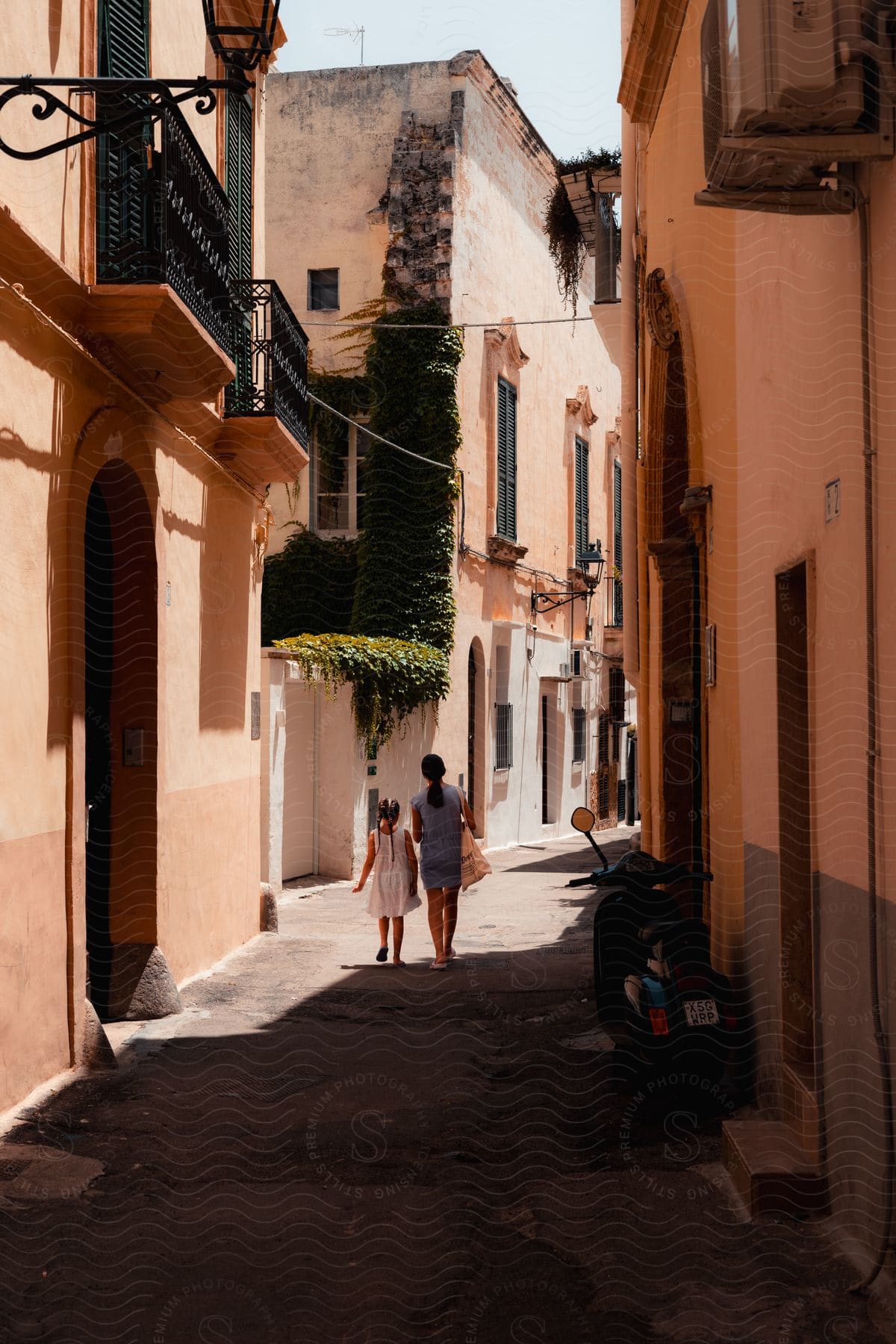 An adult female walking with a young girl in between pink and grey older looking buildings.