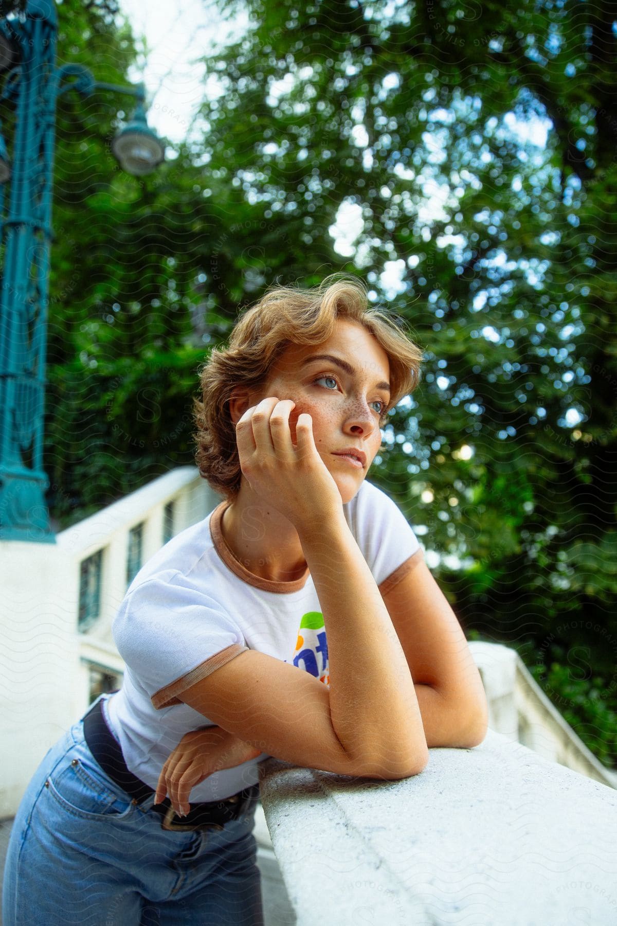 Blonde woman with light eyes leaning on a white balustrade with trees in the background.