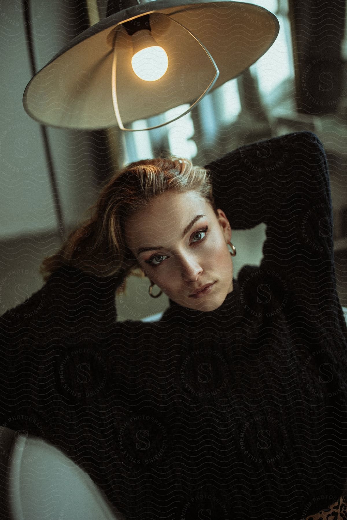 Bathed in the warm glow of a hanging ceiling light, a blonde woman in a black long-sleeved sweater sits gracefully in a living room chair.