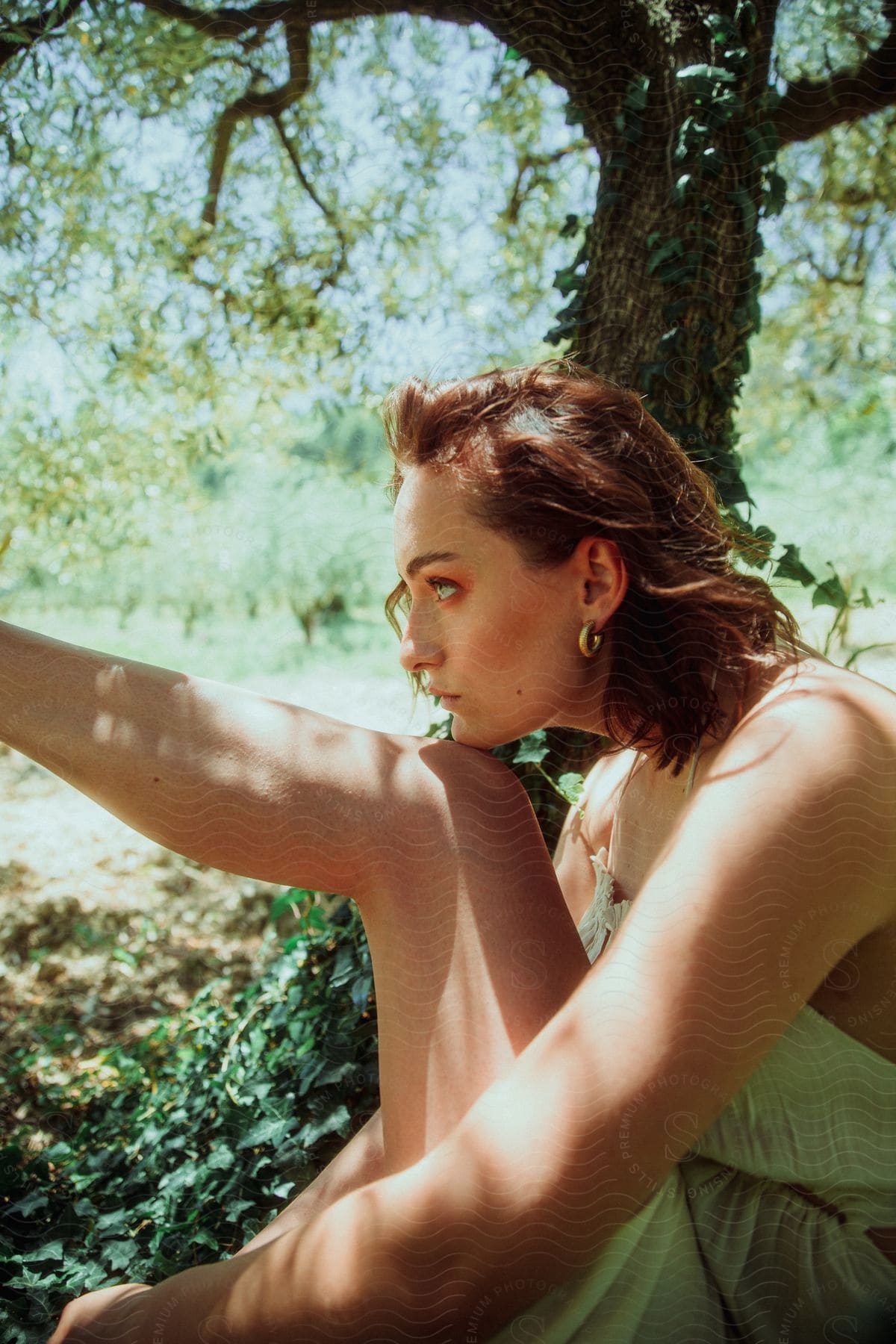 A person with short brown hair sits outdoors in a natural setting, wearing a sleeveless outfit.