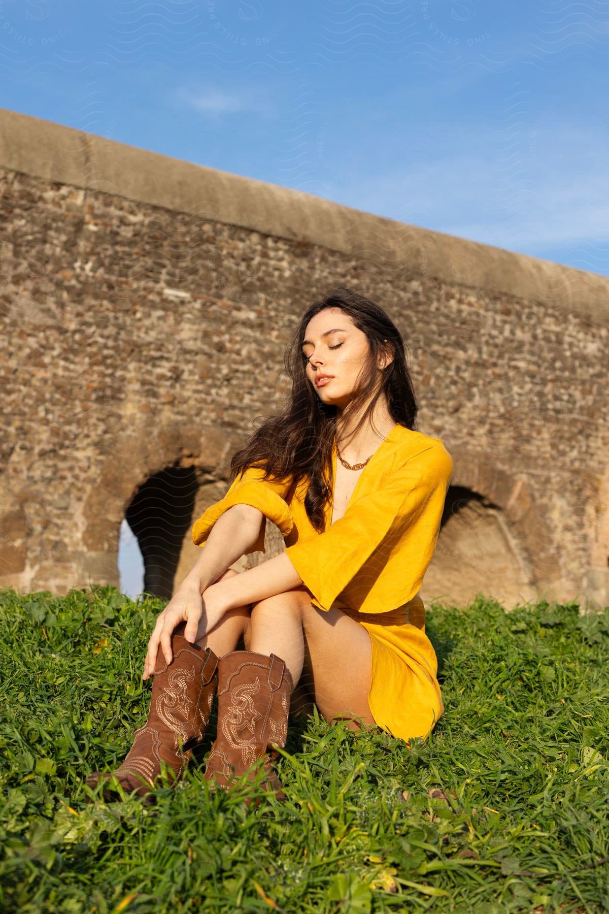 a woman with dark wavy hair, wearing a yellow dress and a pair of black boots, sitting on a green lawn in front of a castle wall.