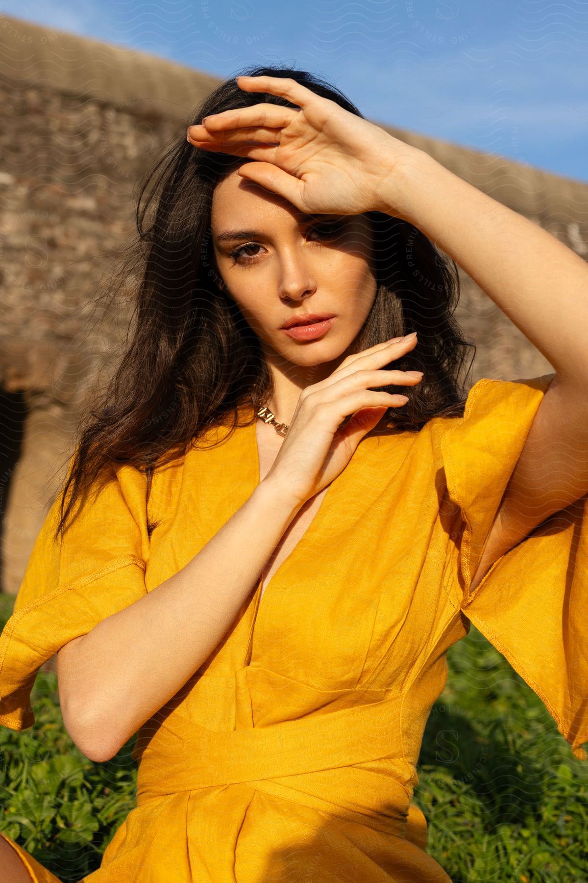 A young woman wearing a yellow dress has one hand on her forehead blocking the sunlight on her face