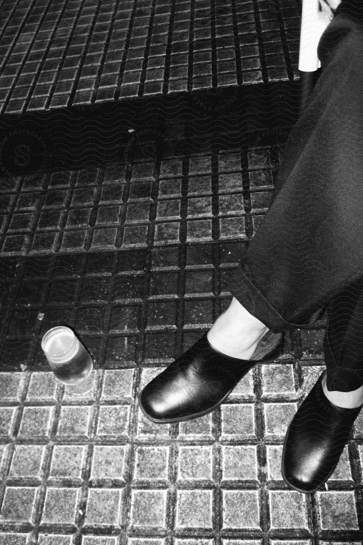 Pair of feet wearing black leather shoes with a clear plastic bottle on the floor beside them.