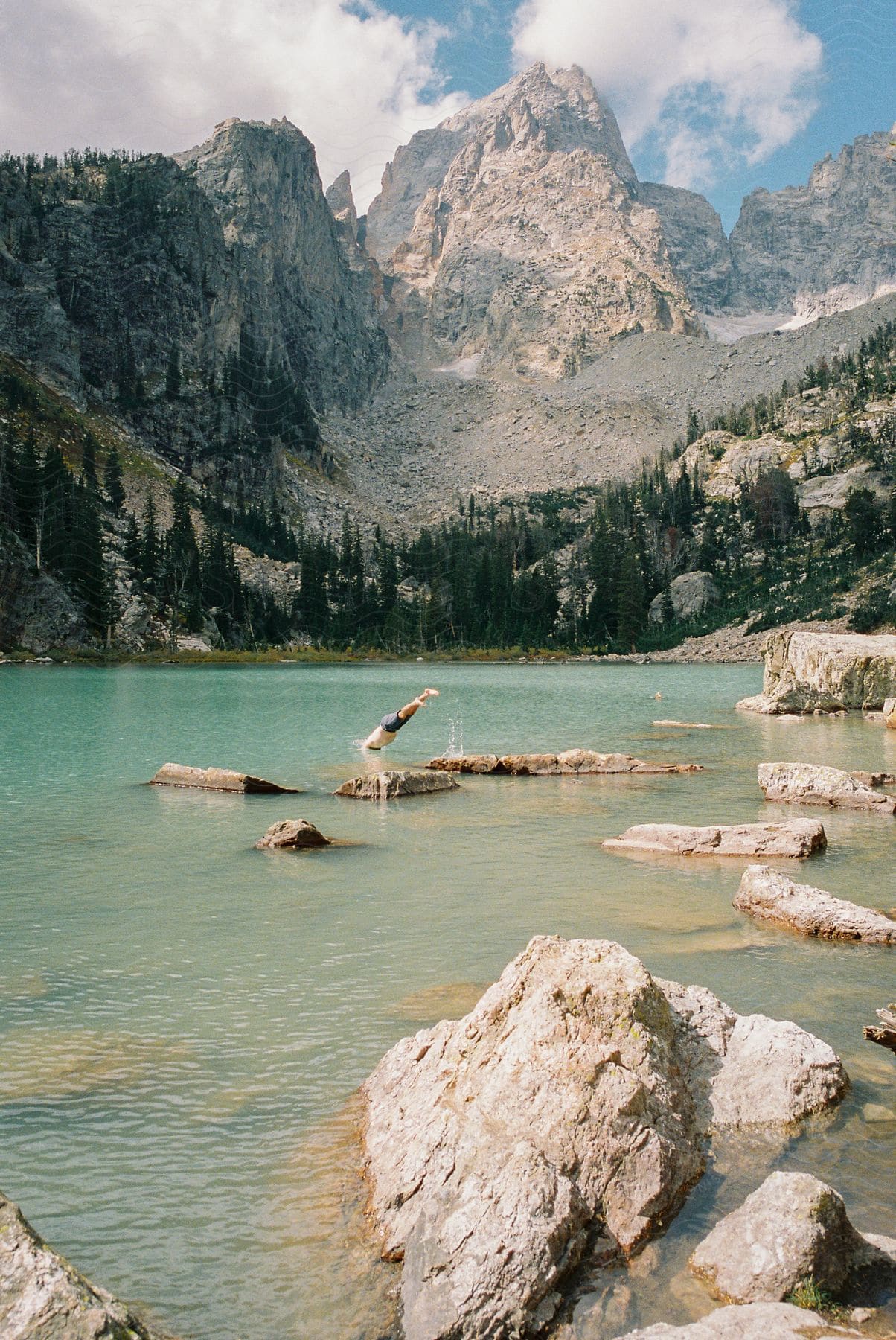 A male diving into water that is near a large mountain range and many rocks in the water.