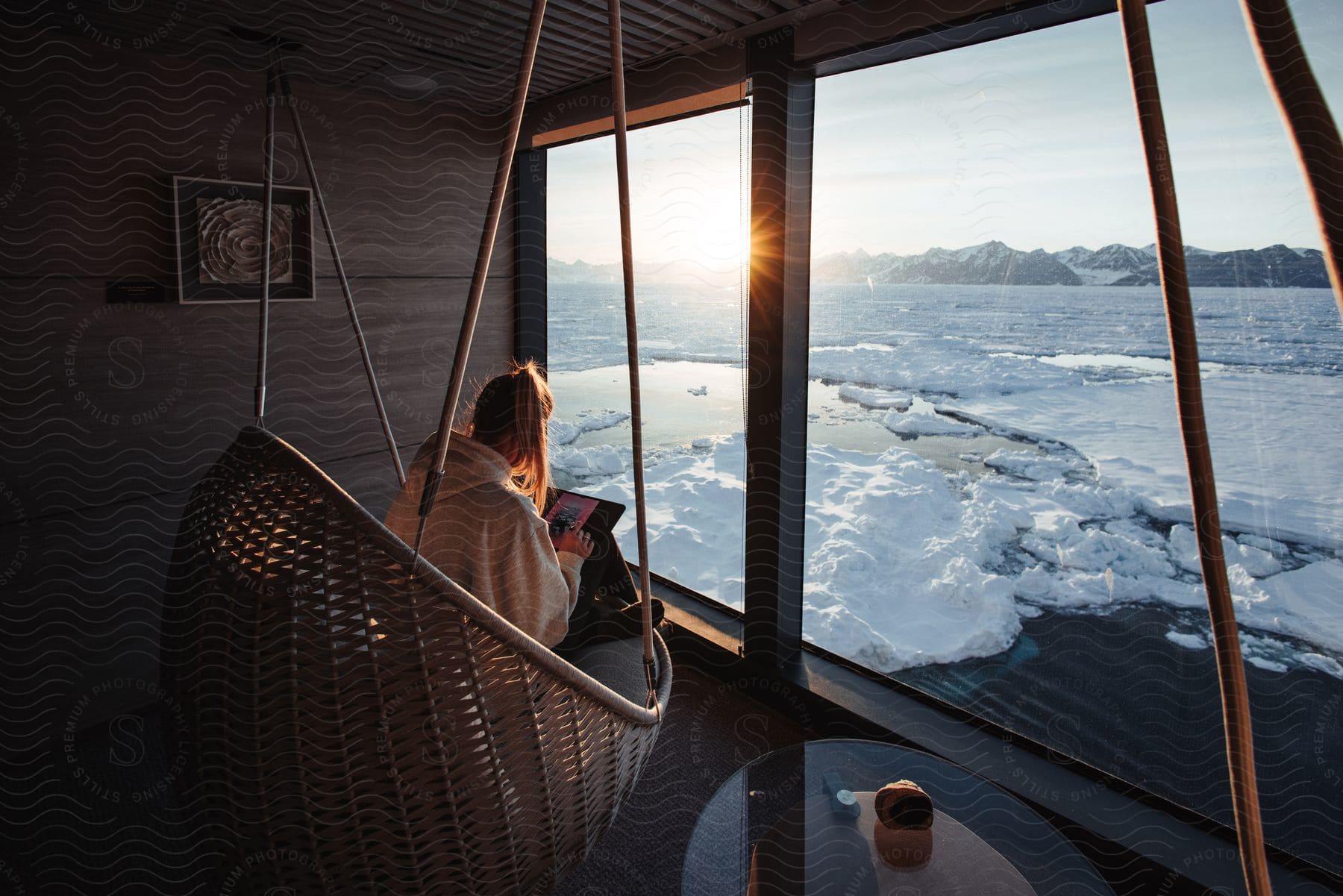 Woman sitting with her back on a hanging chair and using her tablet overlooking a vast landscape of ice and water with mountains in the background during sunset.