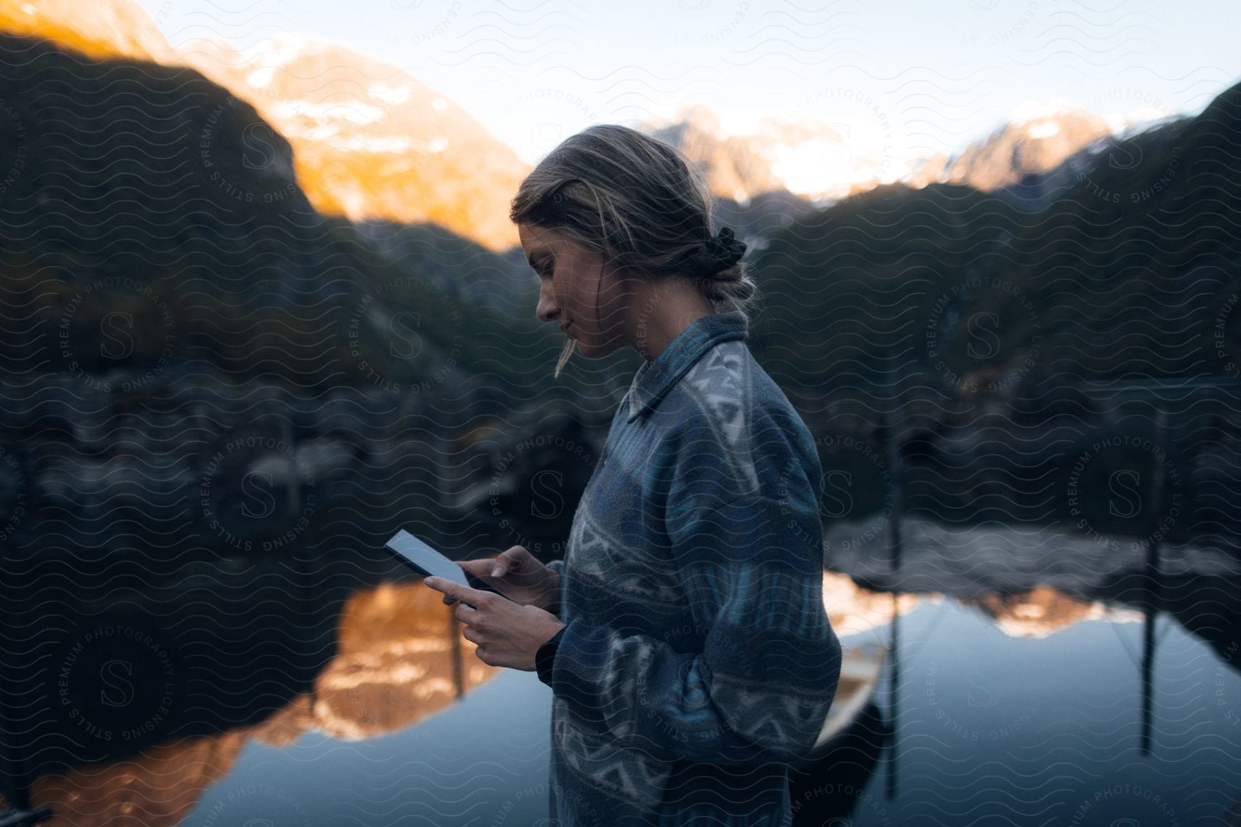 A woman wearing a sweater is looking at her cell phone screen and behind her there is a lake reflecting the surrounding landscape